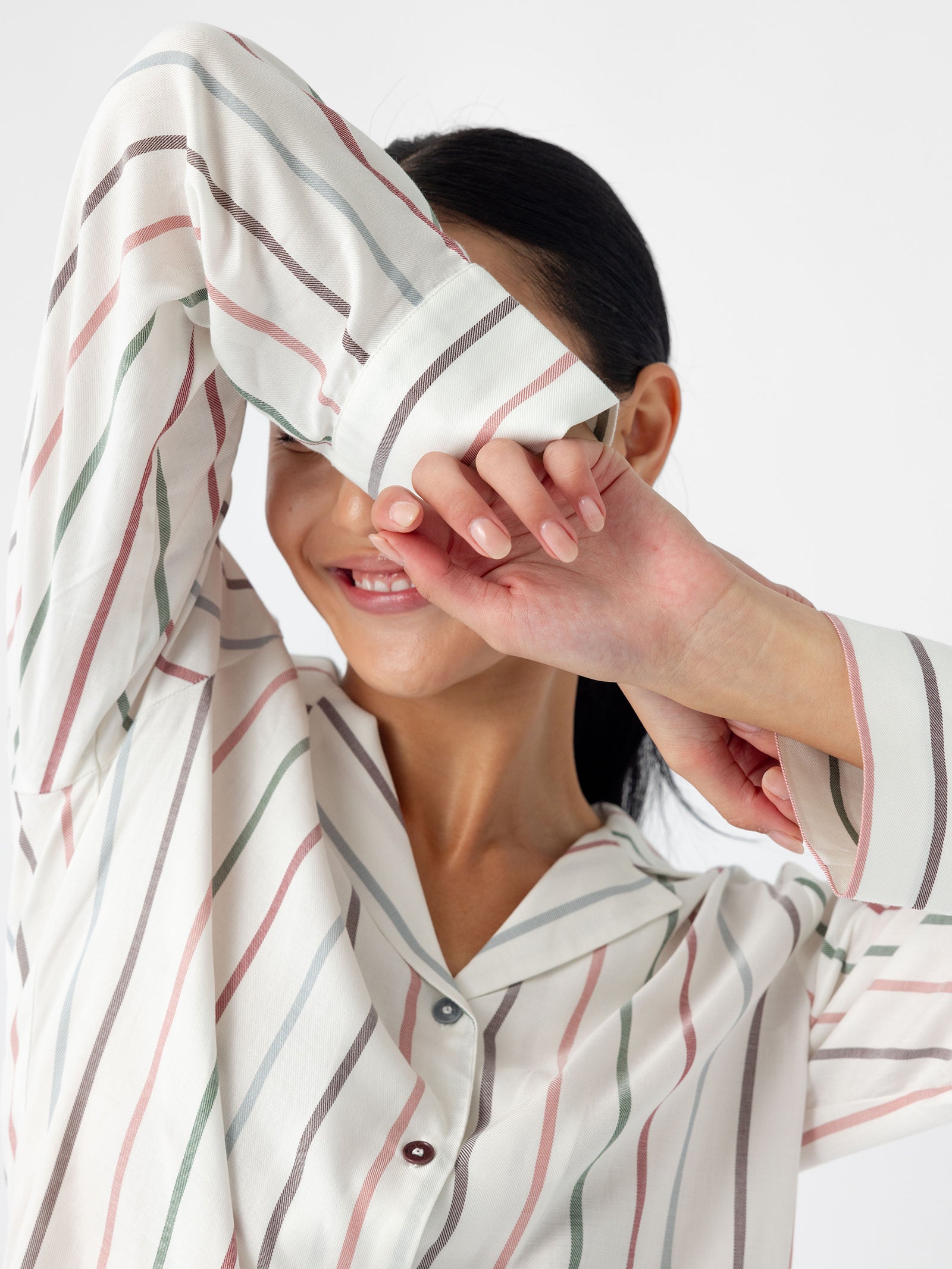 A person with dark hair is smiling and playfully covering their face with their forearm while wearing the Women's Soft Woven Long Sleeve Pajama Top from Cozy Earth. This striped top features pastel colors on a white background. Their nails are neatly manicured, set against a plain white background. 