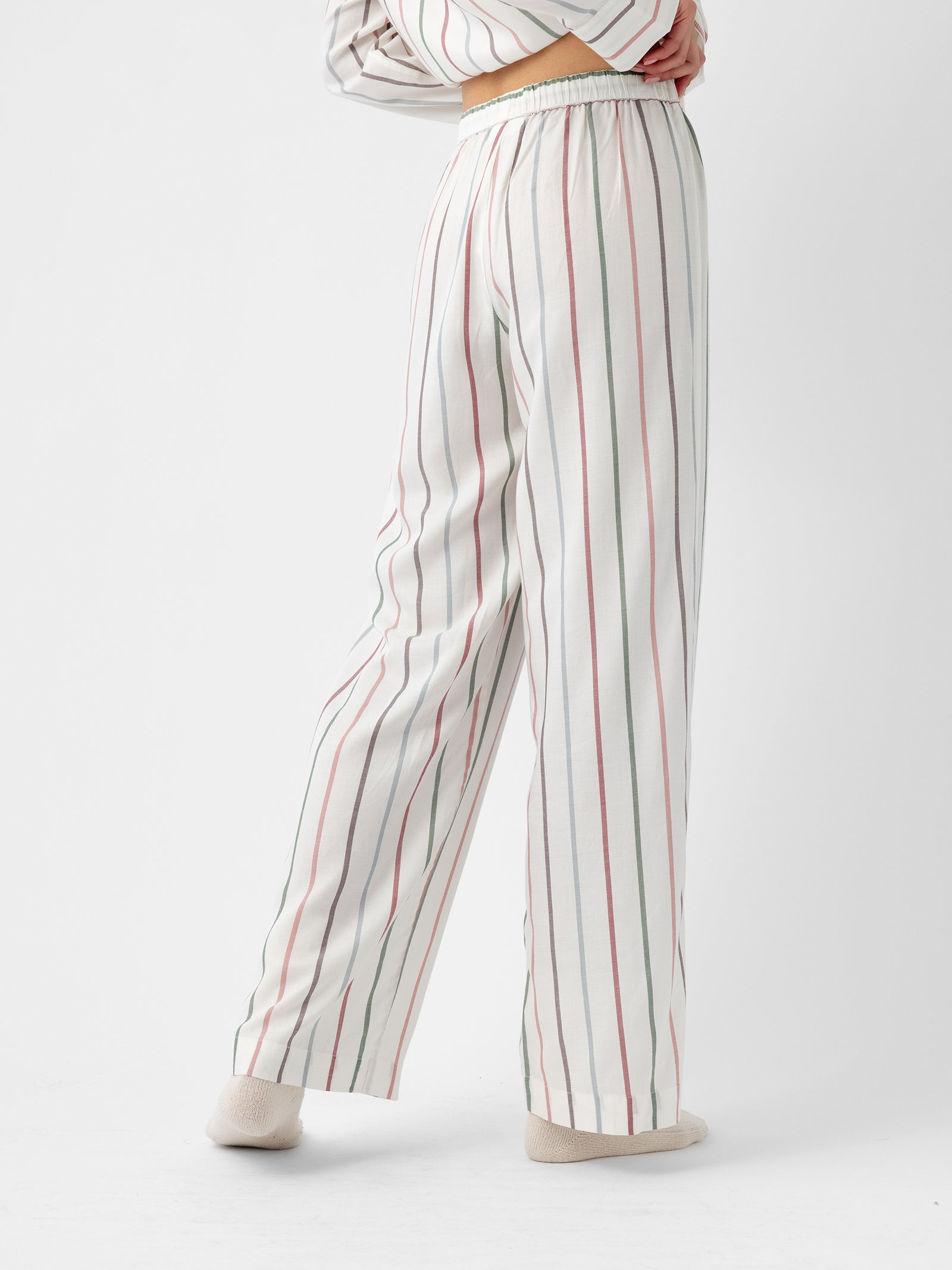 A person is wearing the Women's Soft Woven Pajama Pant from Cozy Earth, featuring cream-colored fabric with vertical stripes in red and green. The pants are styled with a matching long-sleeve top, and the person stands with one leg slightly crossed over the other against a plain white background. 