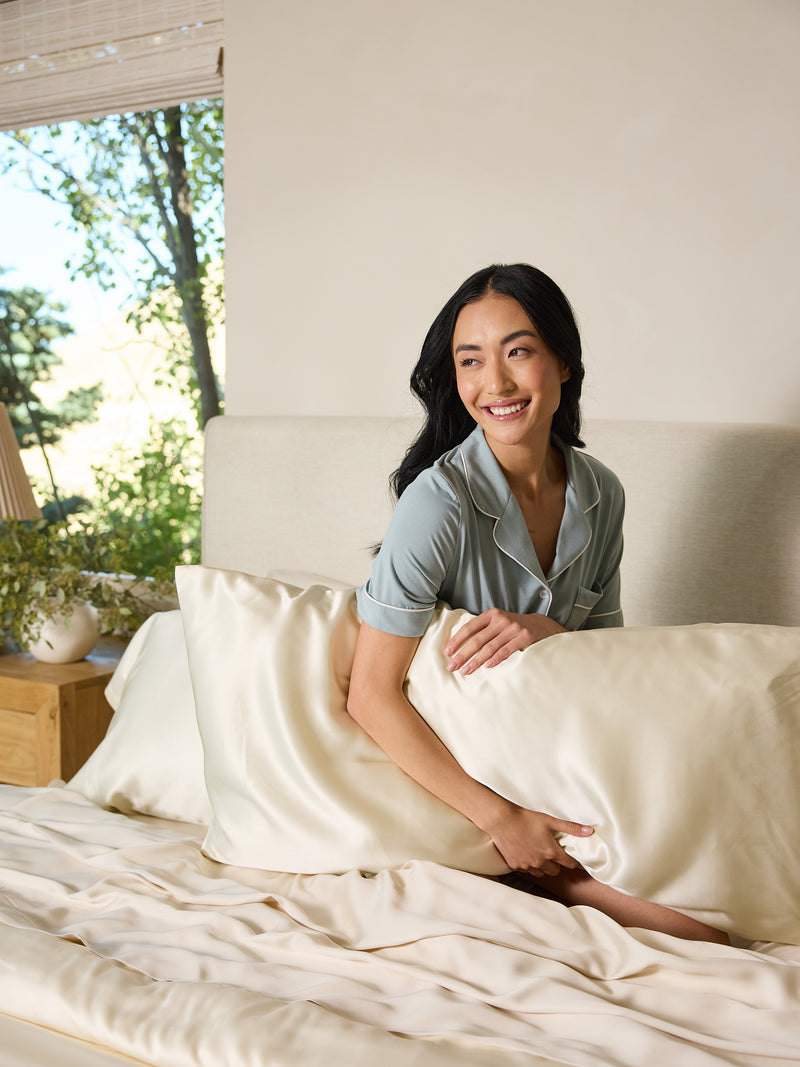 A woman with long dark hair smiles while sitting on a bed, wearing light blue pajamas and holding a large Cozy Earth Silk Pillowcase. Sunlight streams into the bright room through a window, and a wooden nightstand with a plant is nearby.