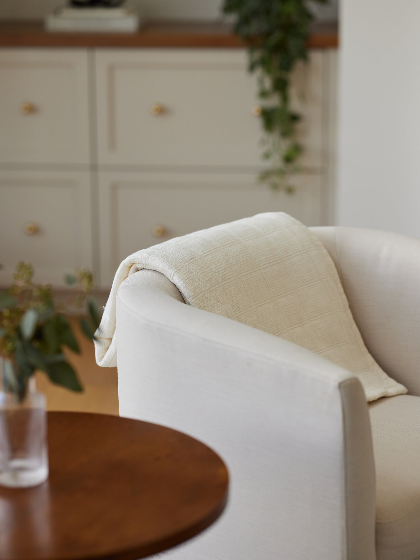 A cozy white armchair with a Waffle Windowpane Blanket by Cozy Earth draped over the backrest sits in a softly lit room. In the foreground, there is a wooden round table with a vase containing greenery. The background features cabinets with gold knobs and a hanging green vine plant. 