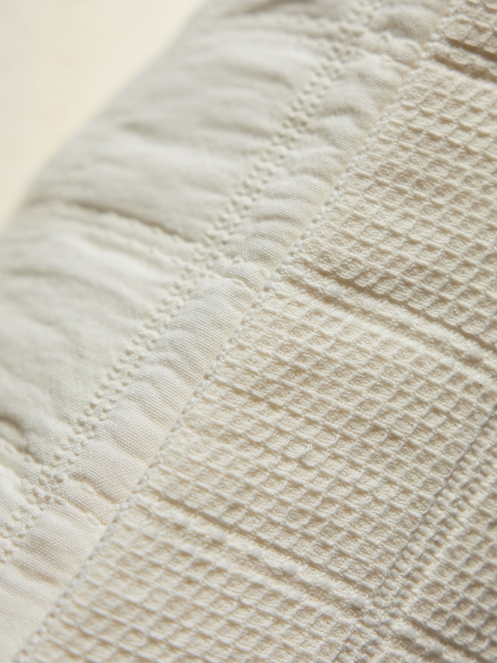 Close-up shot of the Cozy Earth Waffle Windowpane Pillow, featuring its intricate textured fabric with vertical stripes. The pillow showcases both quilted and waffle weave patterns in a subtle off-white hue. The detailed stitching and varied textures create a rich, visually appealing design. 