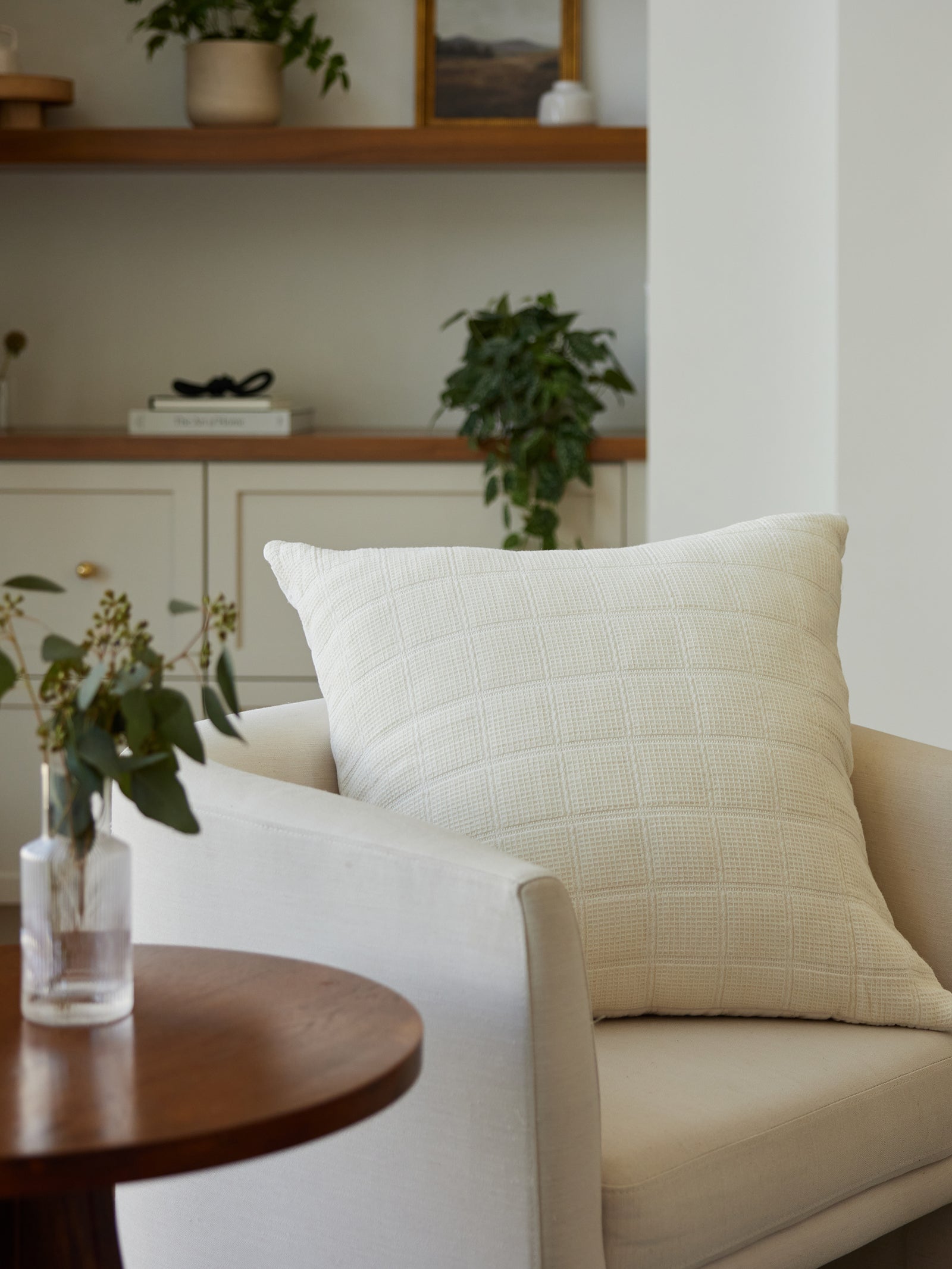 A cozy living room scene features a Waffle Windowpane Pillow by Cozy Earth on a light-colored armchair. In the foreground, a small wooden table holds a clear vase with green foliage. In the background, a beige shelf displays plants and books, enhancing the room's calm ambiance. 