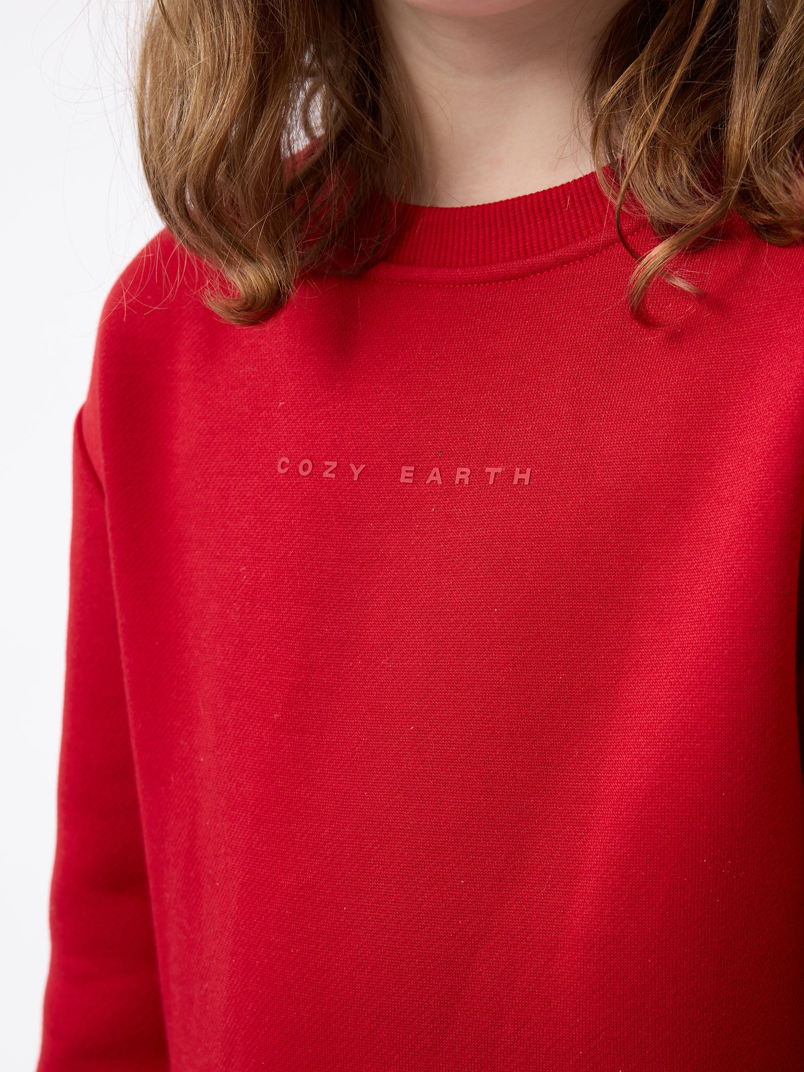 A person with shoulder-length hair wearing a red Kid's CityScape Crewneck by Cozy Earth. The background is plain white. 