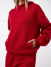 A person wearing a red Women's CityScape Hoodie by Cozy Earth, featuring "Soft Earth" printed on it. The hoodie includes a front pocket, where the person's hands are comfortably tucked. The ensemble is completed with matching red sweatpants against a plain white background. |Color:Crimson