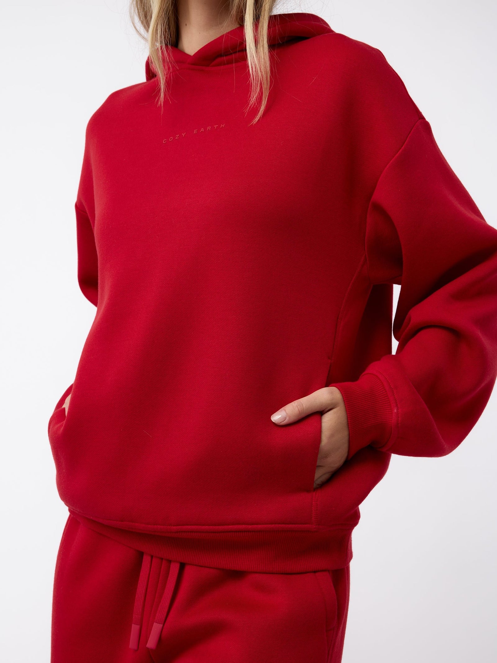 A person wearing a red Women's CityScape Hoodie by Cozy Earth, featuring "Soft Earth" printed on it. The hoodie includes a front pocket, where the person's hands are comfortably tucked. The ensemble is completed with matching red sweatpants against a plain white background. 