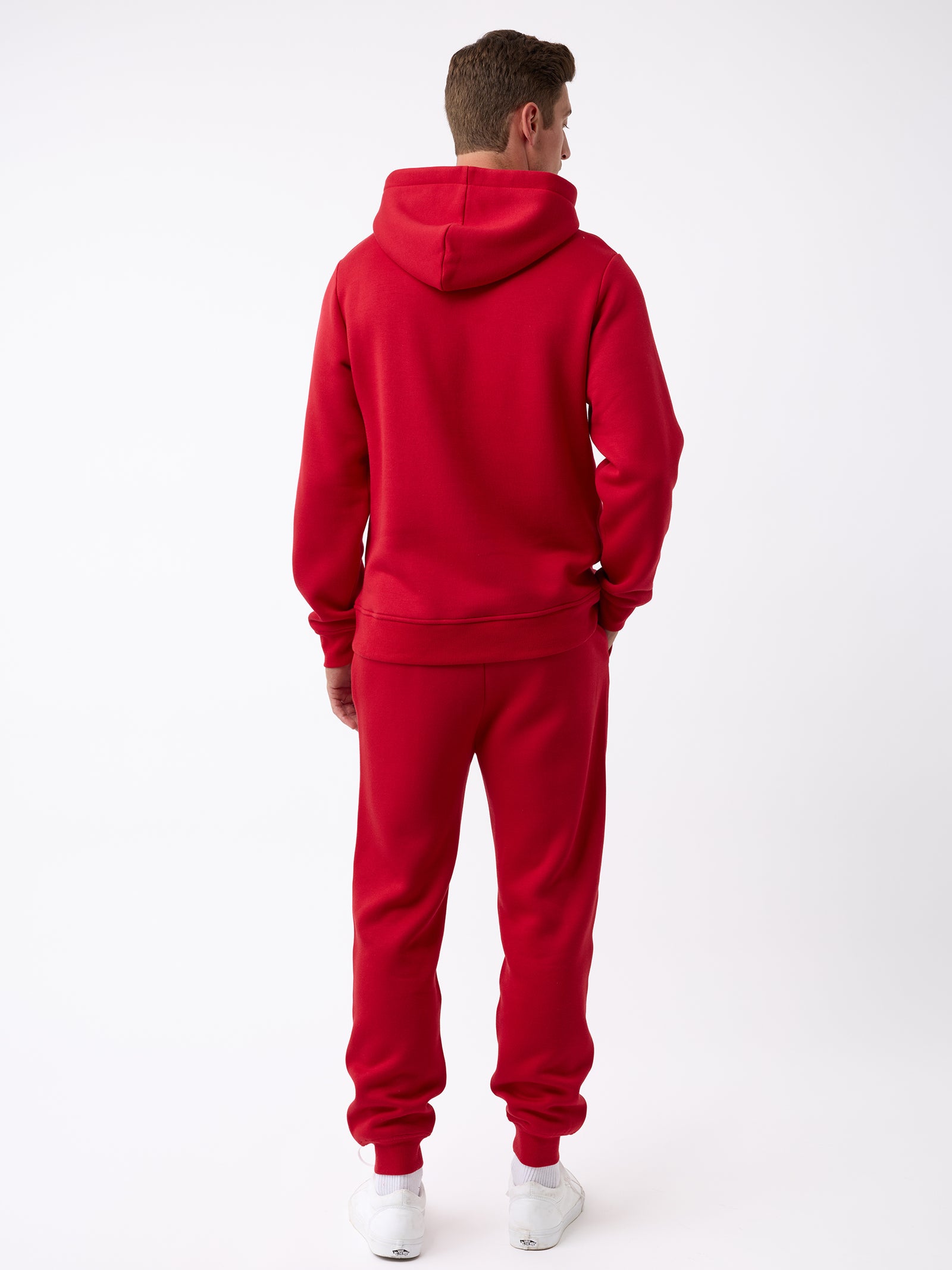 A person is facing away, dressed in Cozy Earth's Men's CityScape Hoodie in a vibrant red and matching red sweatpants. With their hands tucked into the pockets, they are also wearing white sneakers against a plain white backdrop. 