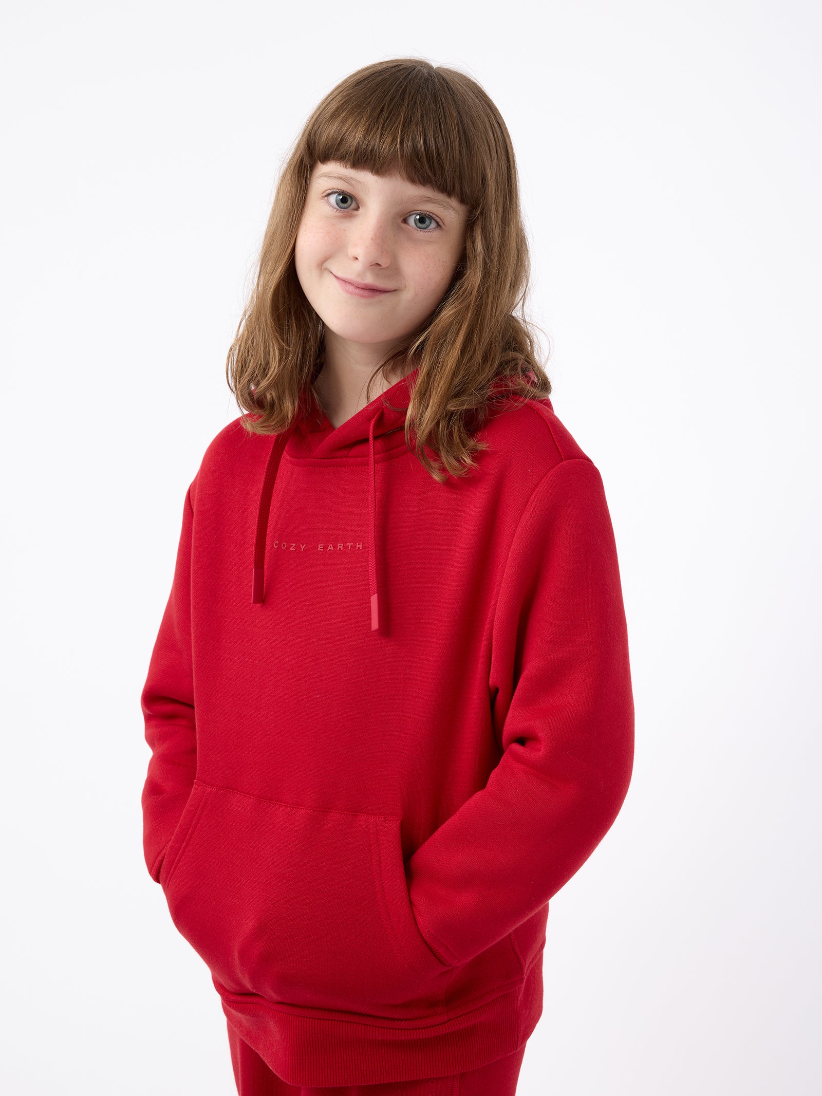 A young girl with shoulder-length brown hair is wearing a Cozy Earth Kid's CityScape Hoodie, featuring her hands in the pockets. She is smiling softly and standing against a plain white background. 