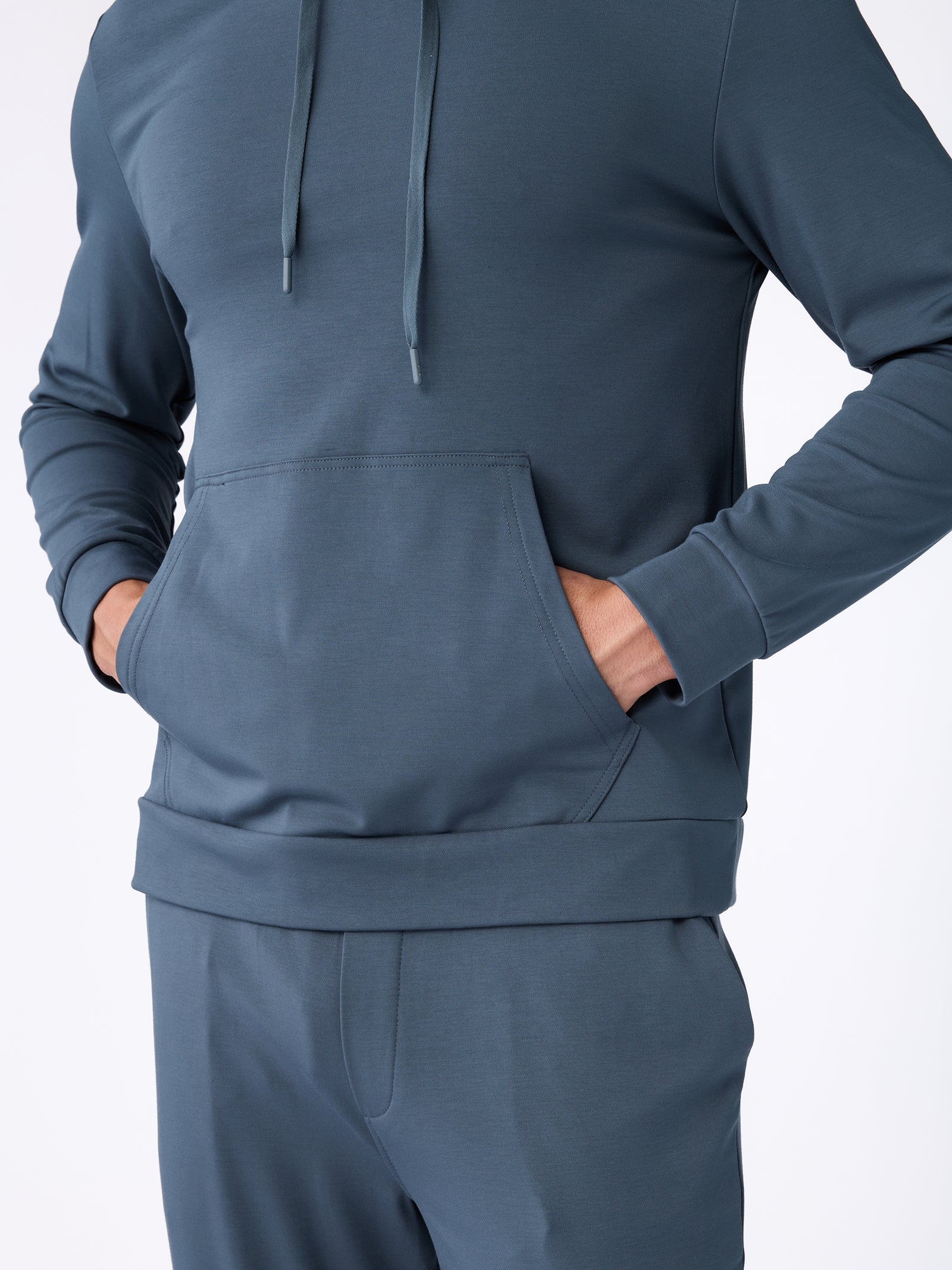 A person is dressed in Cozy Earth's Men's Ultra-Soft Bamboo Hoodie, featuring a matching blue ensemble with hands in the front pocket, exuding a comfortable and casual vibe against a plain background. 