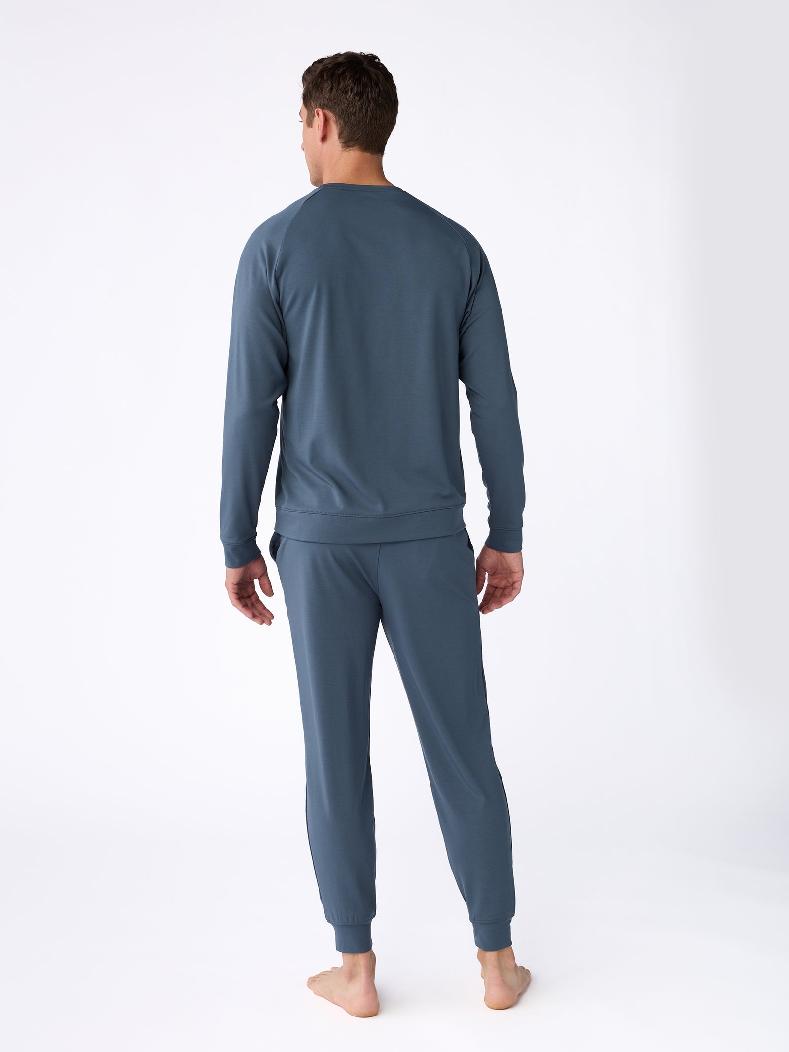 A person wearing a blue Men's Ultra-Soft Bamboo Pullover Crew by Cozy Earth stands barefoot on a white background, facing away from the camera. 