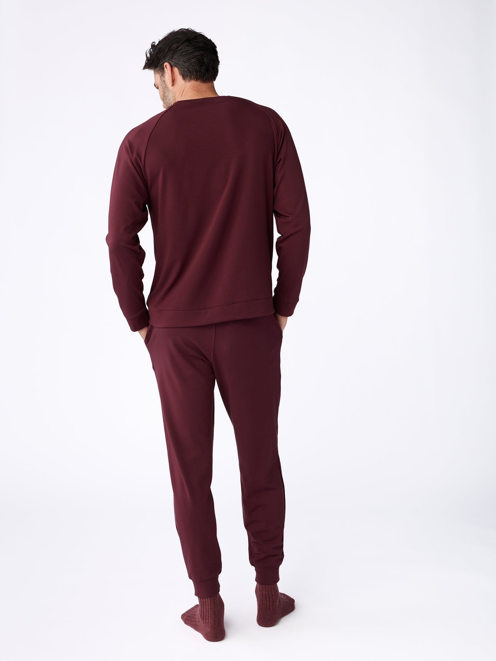 A person stands facing away, wearing the Men's Ultra-Soft Bamboo Pullover Crew from Cozy Earth in maroon, paired with matching pants. They are barefoot, set against a plain white background. 