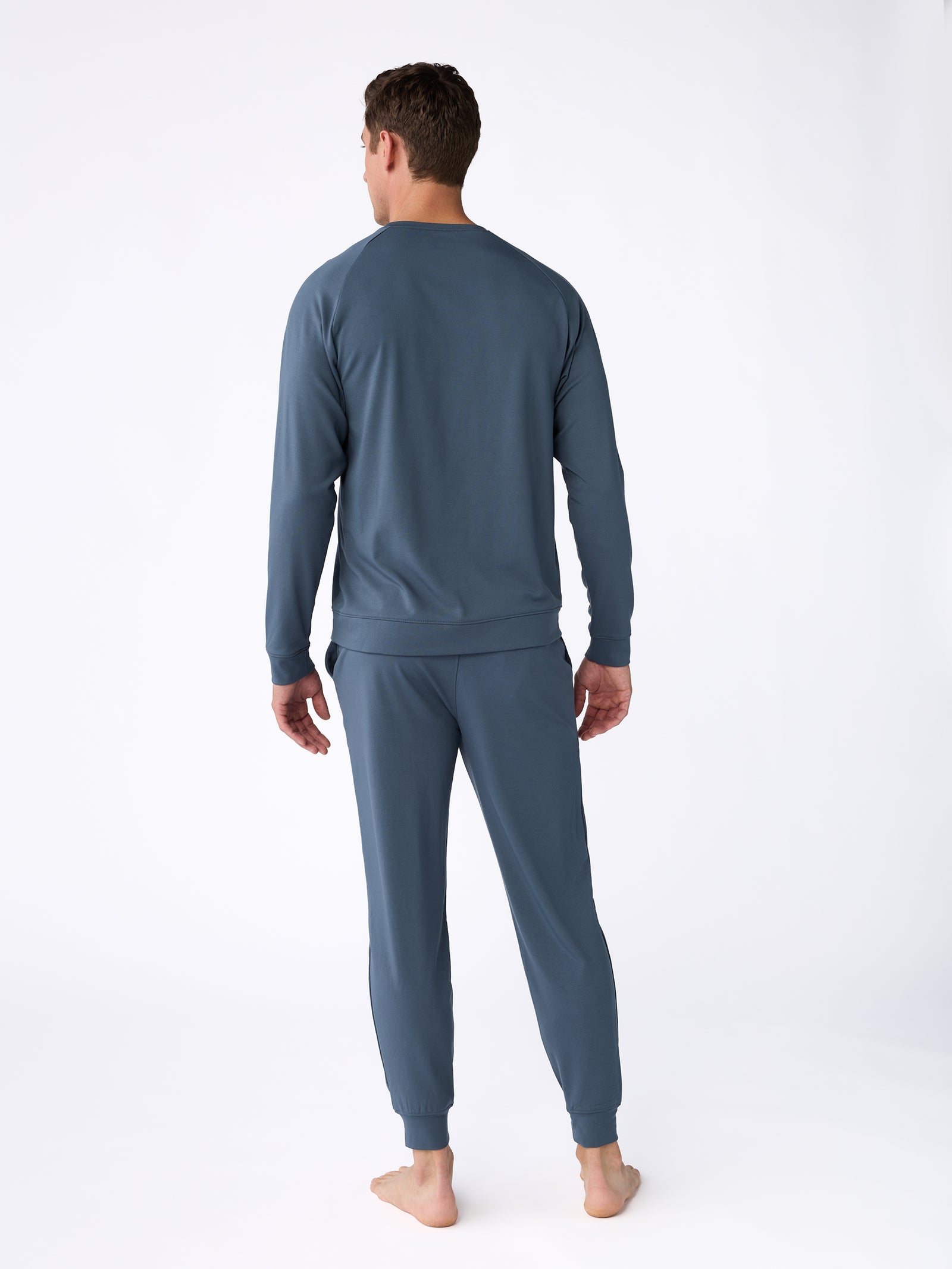 A man stands barefoot with his back facing the camera, wearing the Men's Ultra-Soft Bamboo Pullover Crew by Cozy Earth, a blue long-sleeve top and matching pants. The background is plain white. 