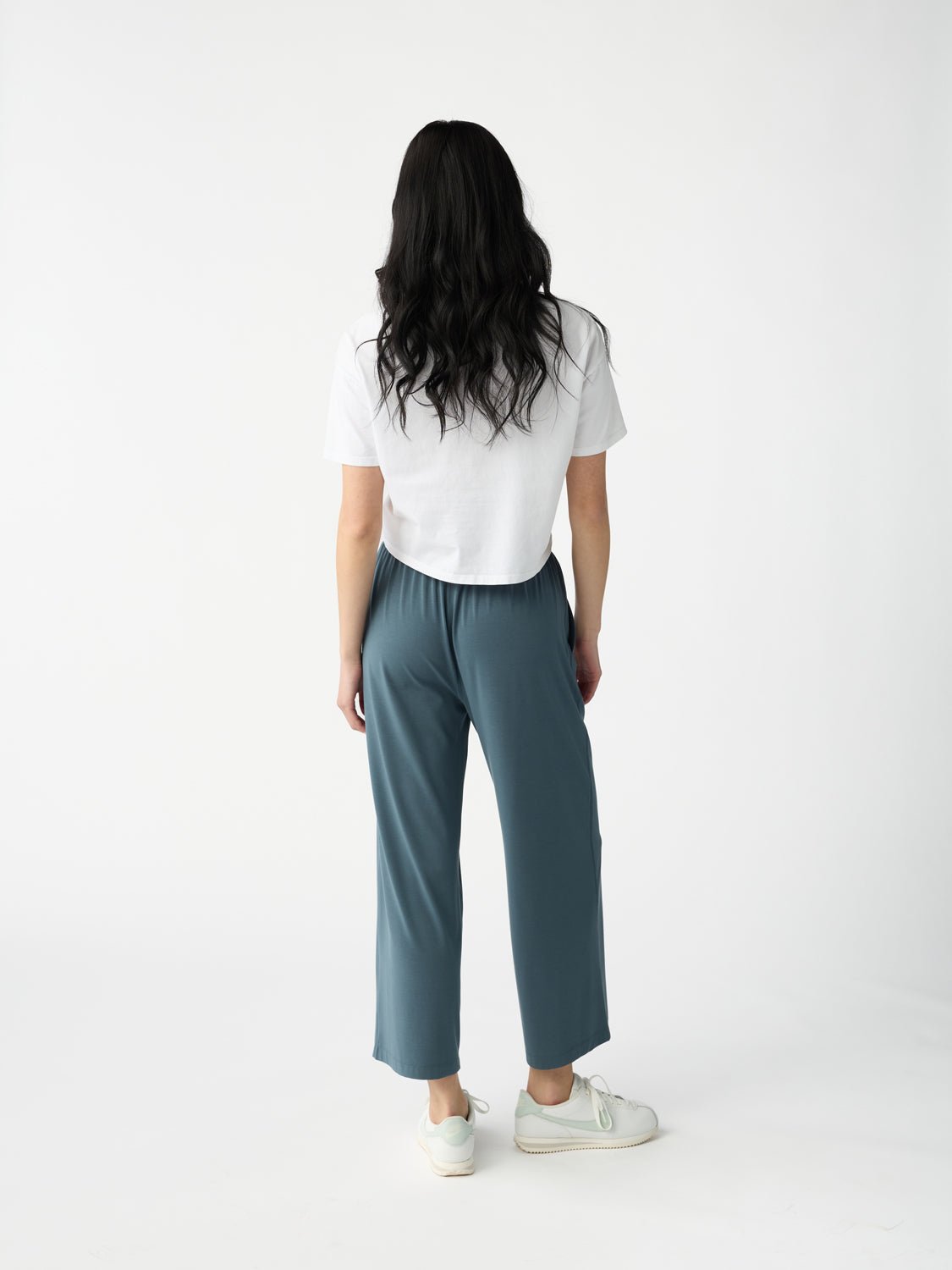 A person with long dark hair stands facing away, wearing a white T-shirt, Cozy Earth Women's Studio Cropped Wide Leg Pant in teal, and white sneakers. The background is plain white. 