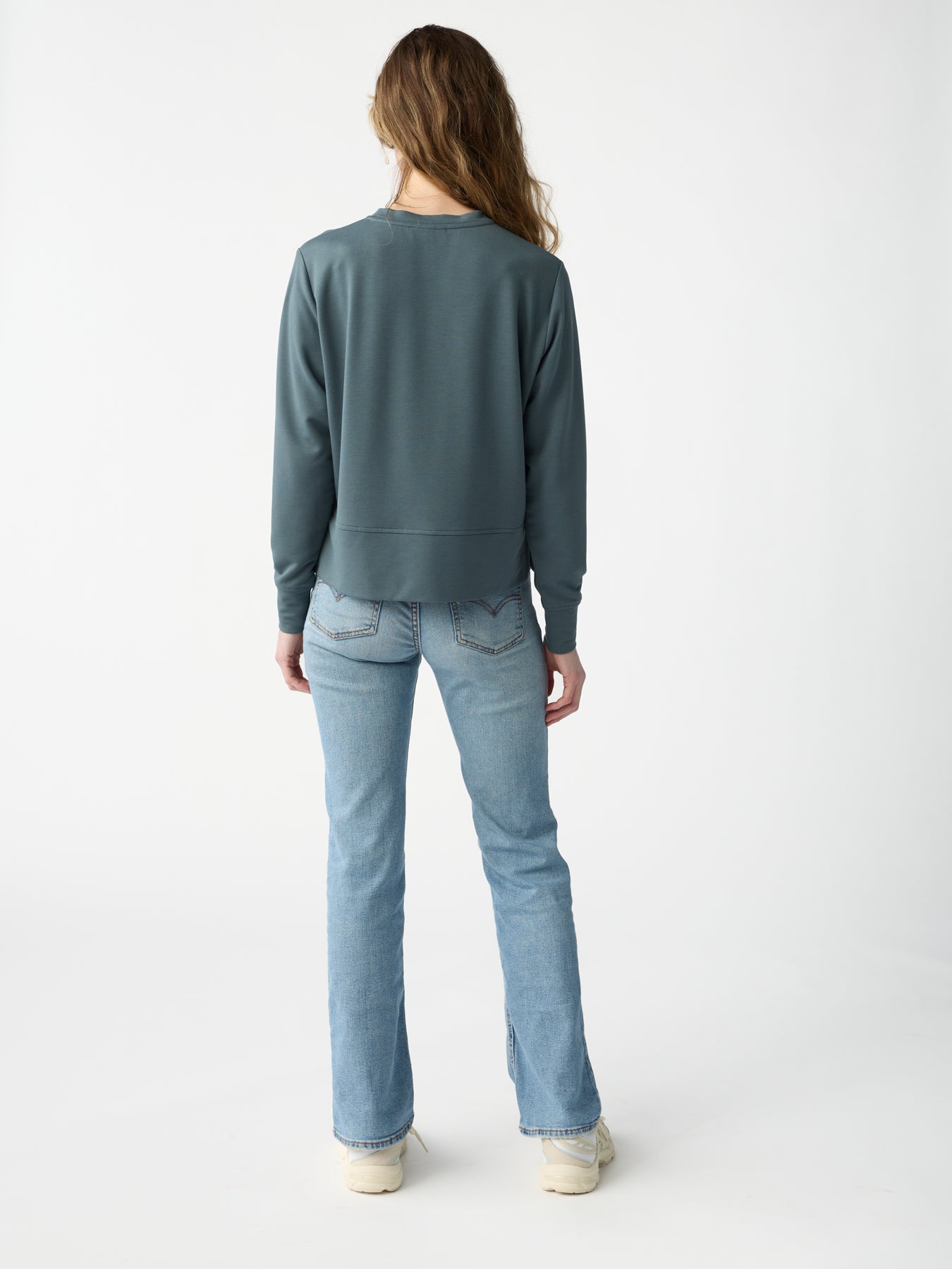 A person with long hair, dressed in a Cozy Earth Women's StudioLite Crewneck sweater in teal and light blue jeans, is standing with their back to the camera against a plain white background, wearing white shoes. 