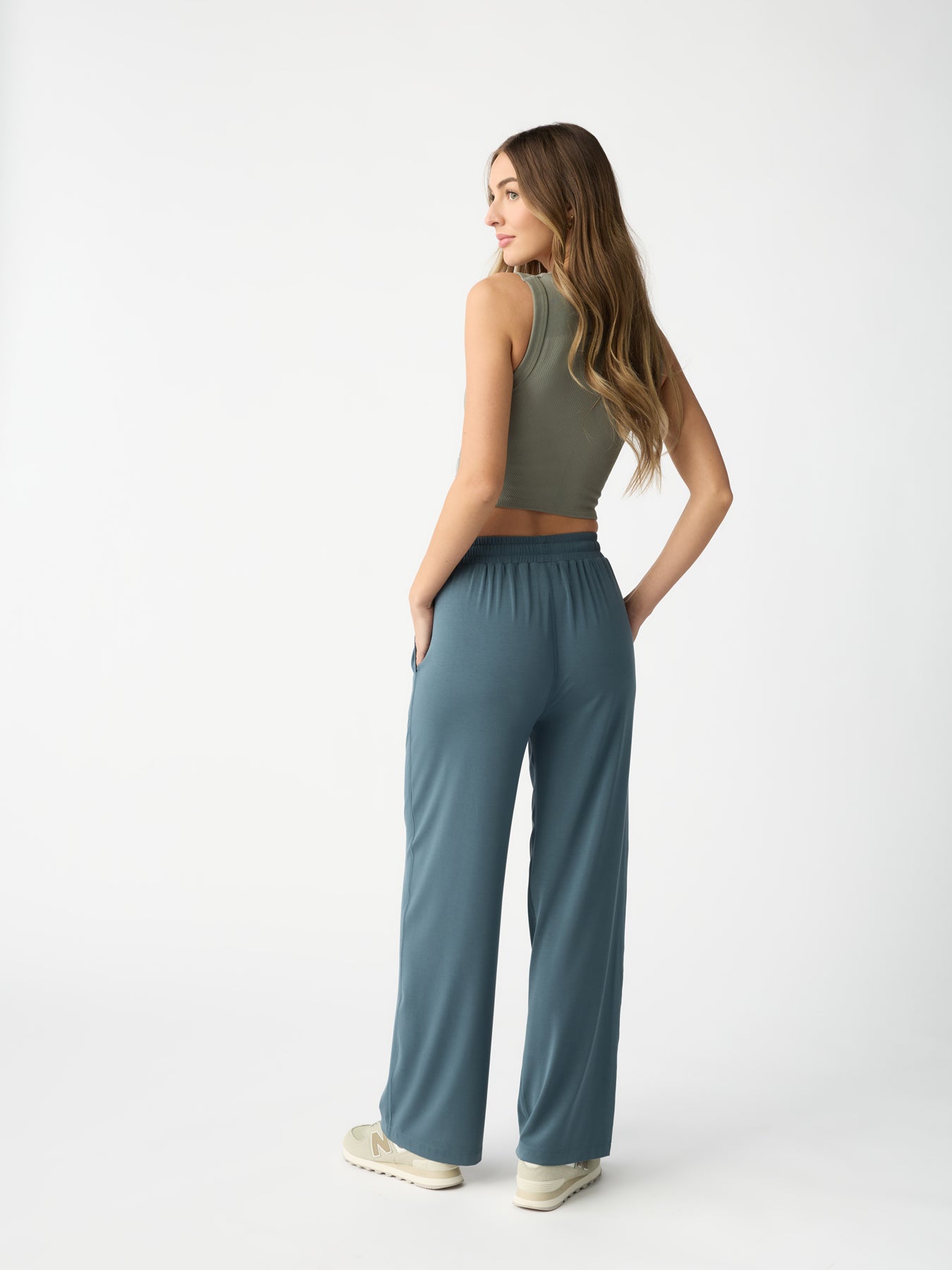 A woman with long hair stands sideways against a plain white background, wearing Cozy Earth's sleeveless olive green top and Women's Studio Wide Leg Pant in blue, paired with sneakers. Her hands are in her pockets. |Color:Dawn