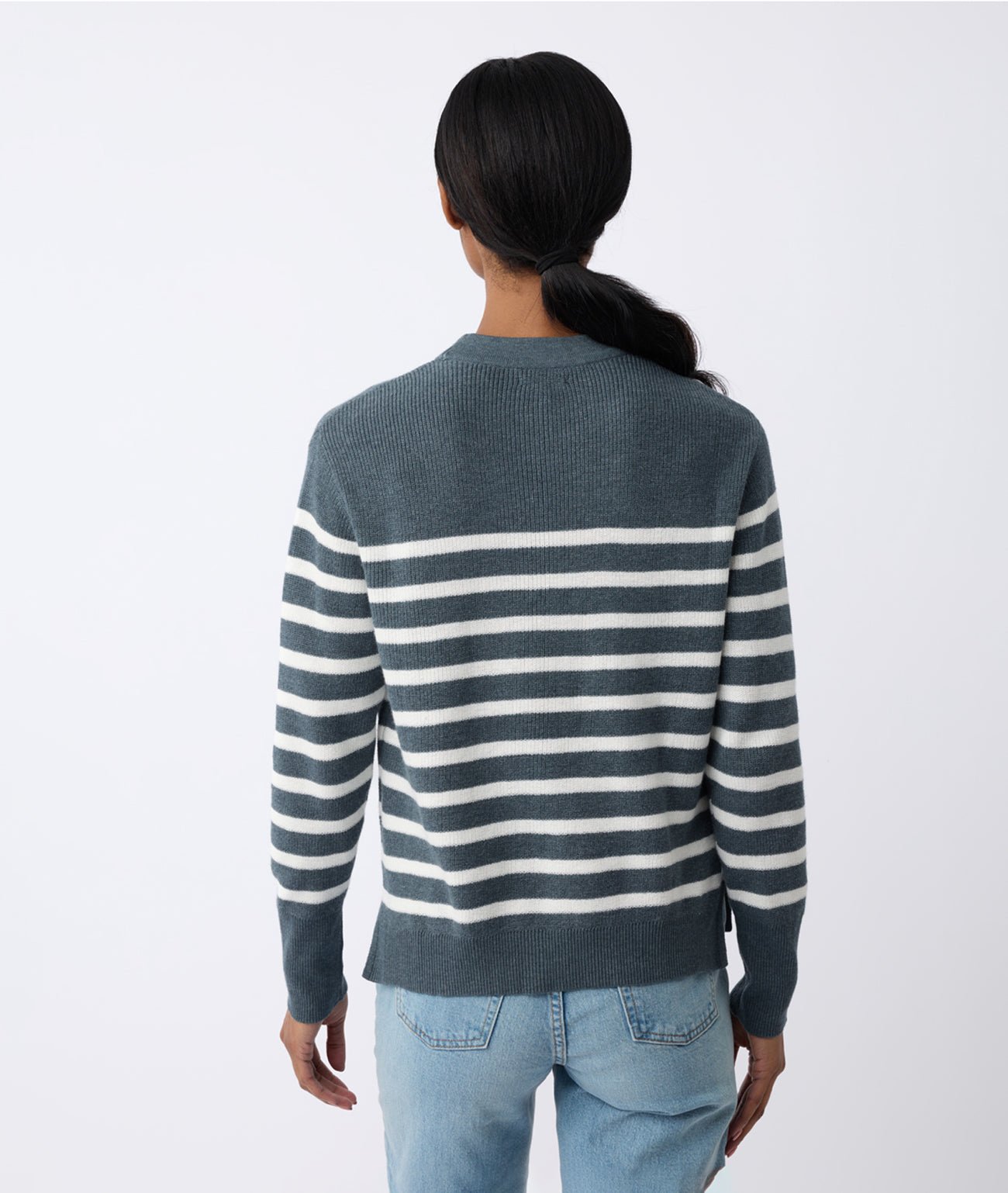 A person with long dark hair is seen from the back, wearing a Cozy Earth Women's Rowan Sweater paired with light blue jeans, standing against a light gray background. 