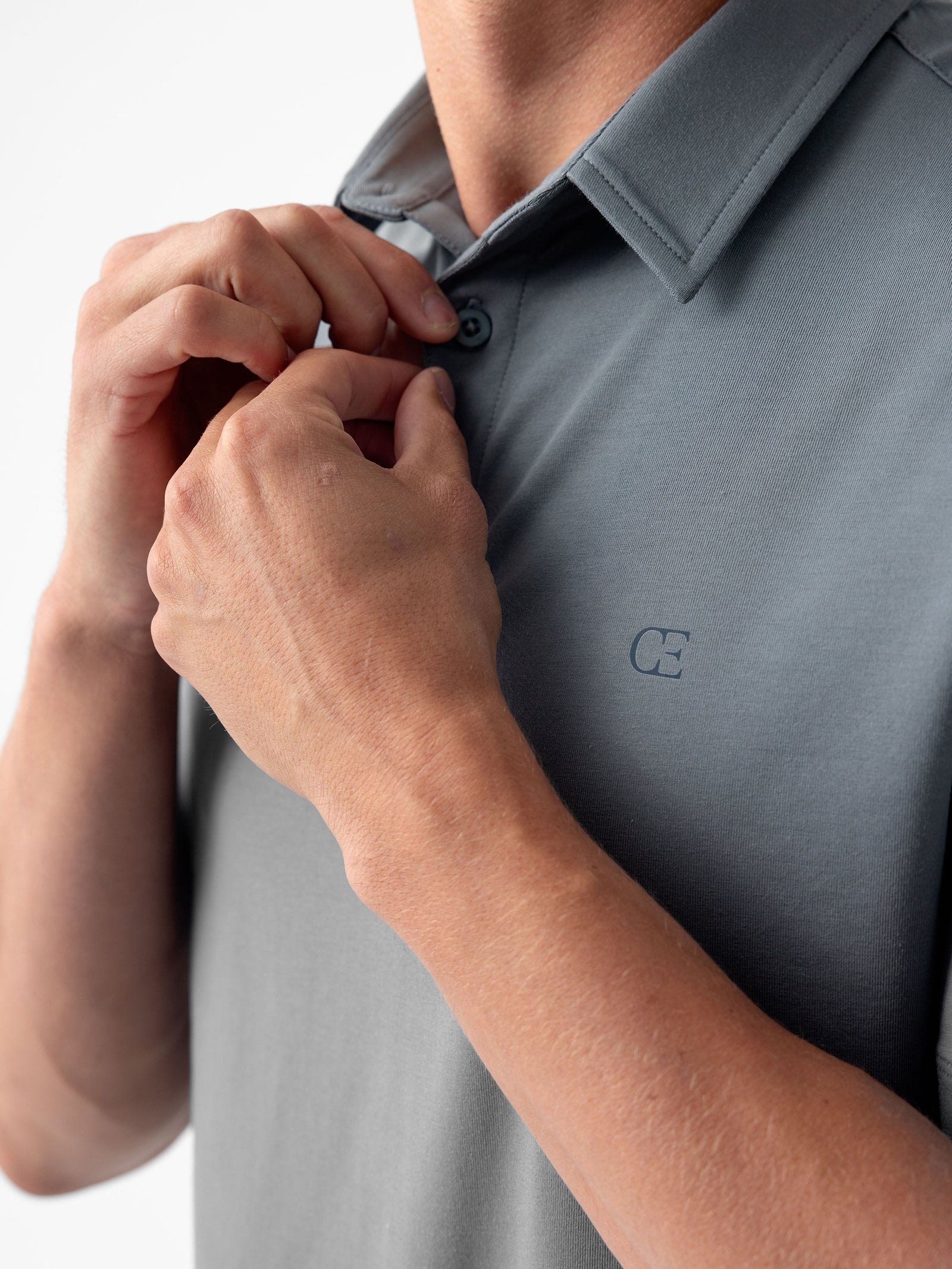 A person buttoning a Cozy Earth Men's Everyday Polo in gray. The polo features a small, embroidered logo on the chest. The focus is on the hands and upper torso against a plain background. 