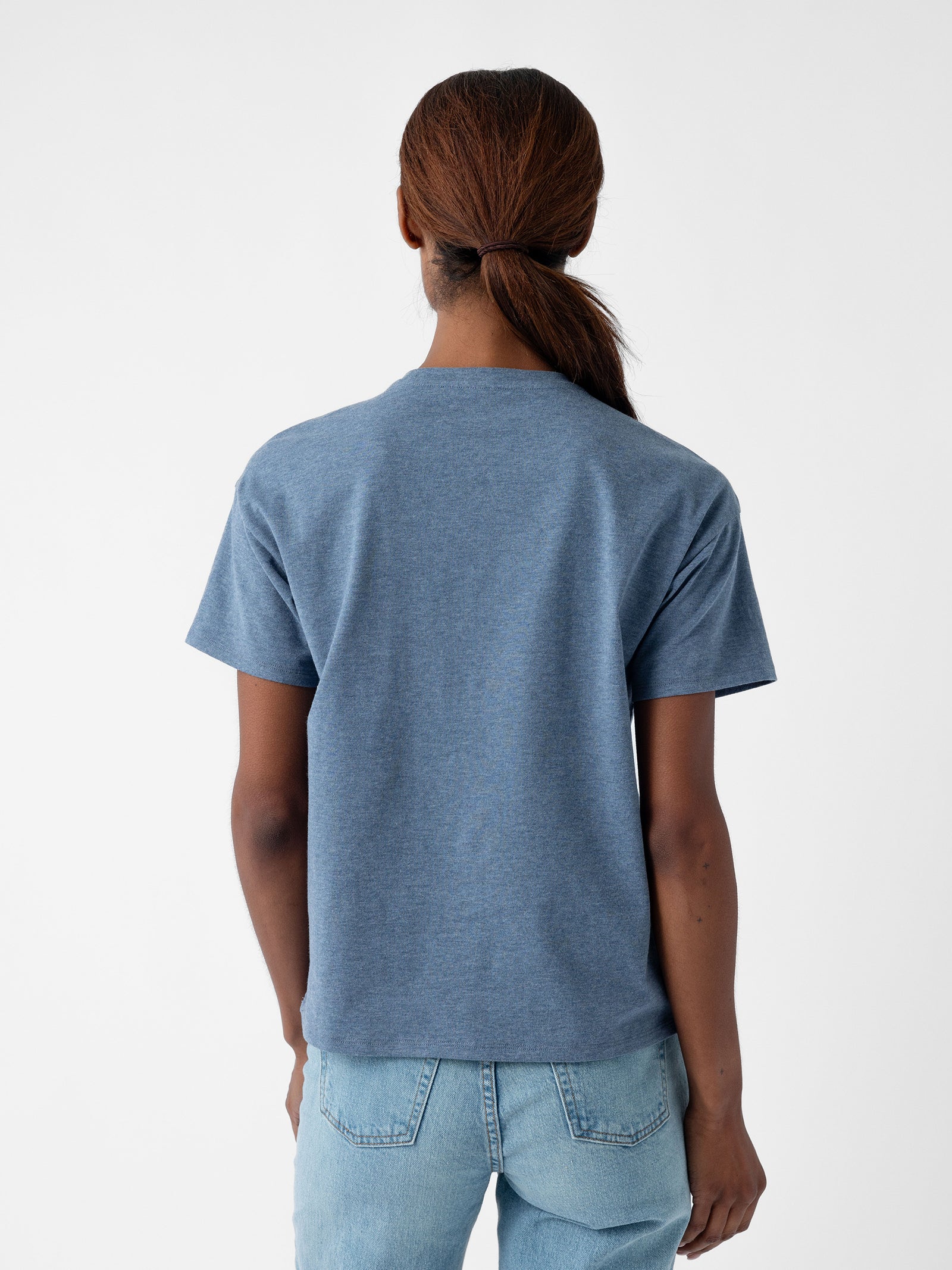 A person with long hair tied in a low ponytail is wearing the Women's All Day Tee by Cozy Earth and light blue jeans. They are standing with their back facing the camera against a plain white background. 