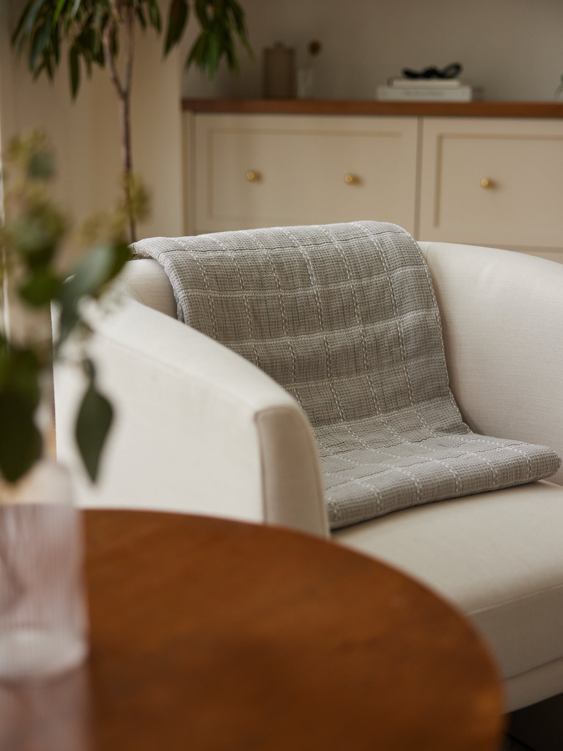 A cozy living room scene featuring a light-colored armchair with the Cozy Earth's Waffle Windowpane Blanket draped over the backrest. In the foreground, there is a round wooden table with a vase of greenery. A dresser with brass knobs is in the background. |Color:Dove Grey
