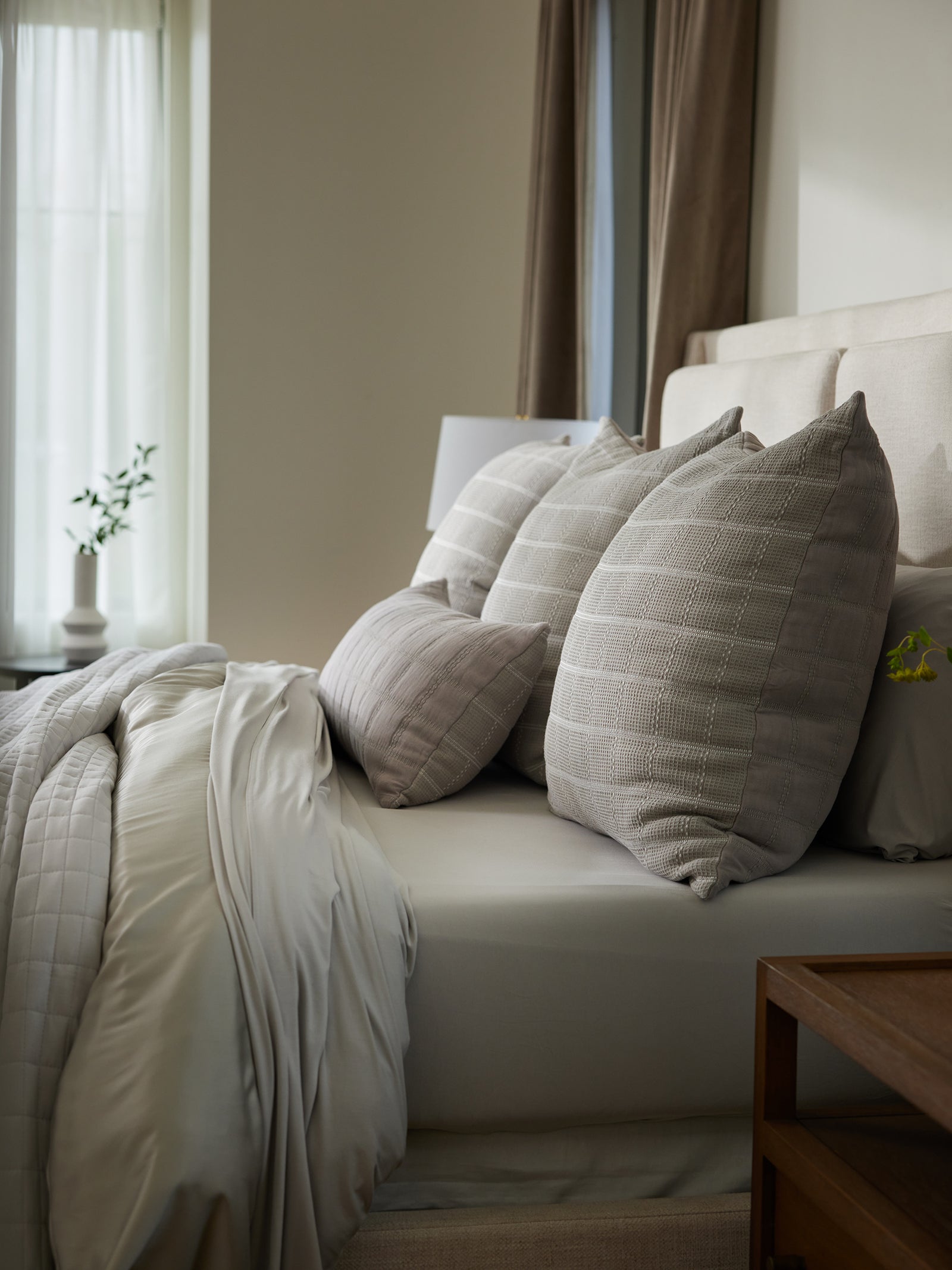 A neatly made bed features beige and light gray bedding, adorned with several textured throw pillows, including the Waffle Windowpane Pillow from Cozy Earth. Natural light streams in from a window, while a small plant in a white vase rests on a bedside table, enhancing the serene and cozy atmosphere. 