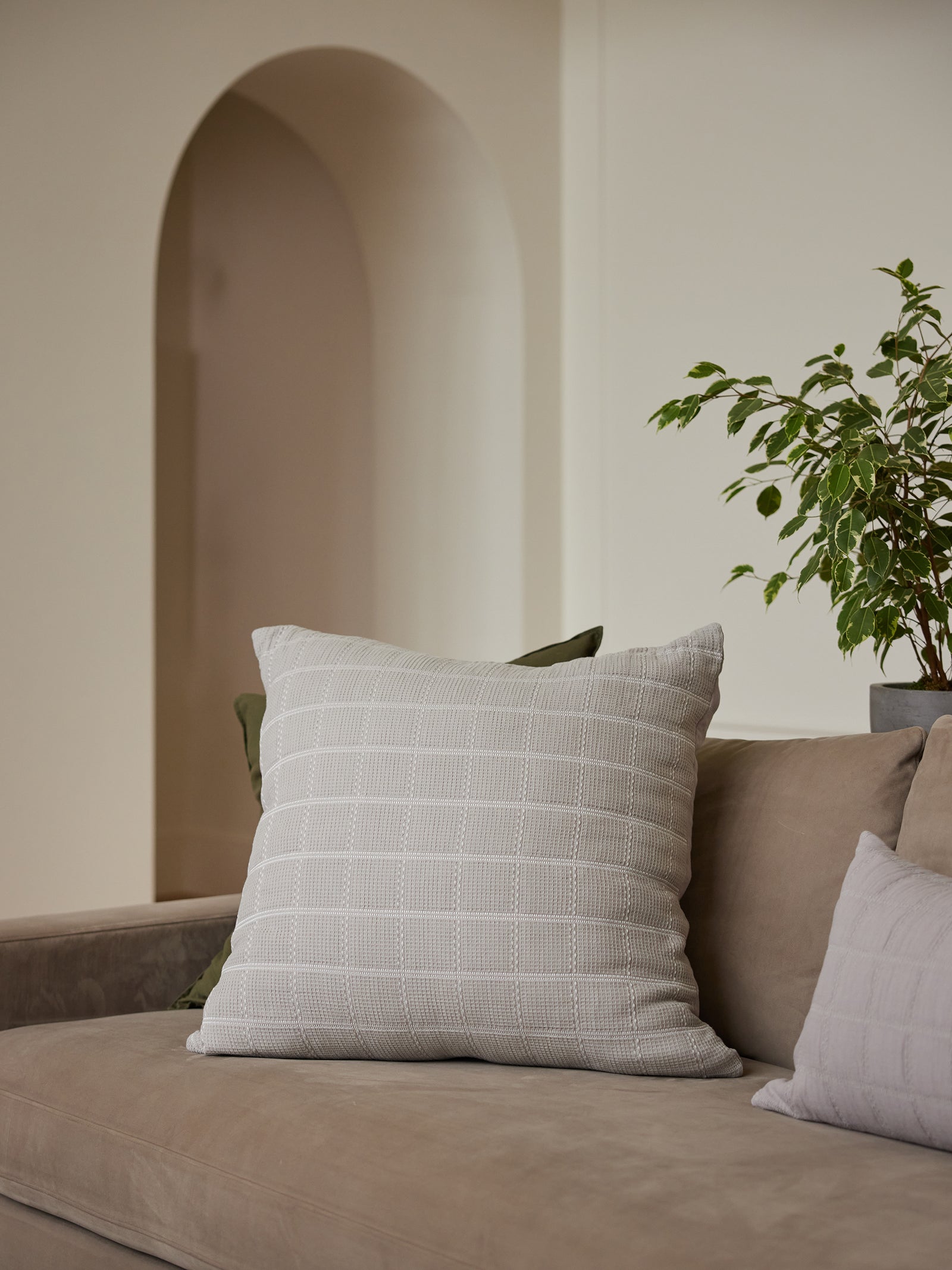 A beige couch adorned with a large, textured Cozy Earth Waffle Windowpane Pillow in white and a smaller grey cushion. To the right, a green plant in a grey pot sits on the arm of the couch. An archway in the background opens up to another part of the room, all warmly lit. 