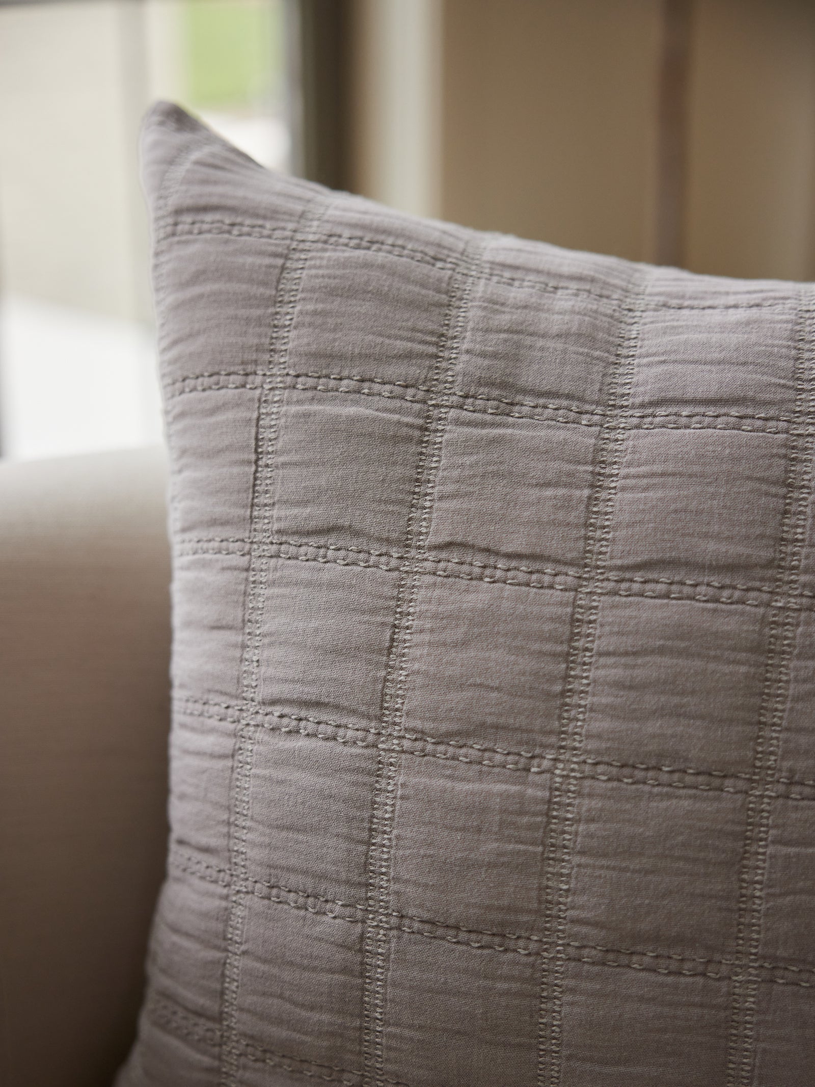 Close-up of the Waffle Windowpane Pillow by Cozy Earth, featuring a light beige hue and a textured grid pattern, resting on a light-colored armchair. The softly blurred background highlights what seems to be a window or natural light source. 