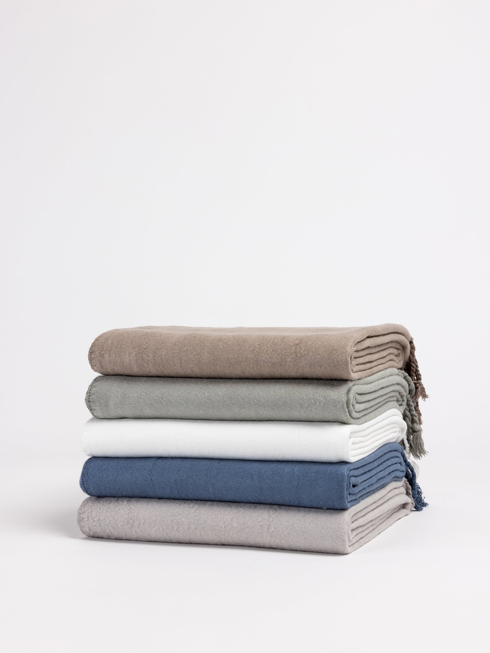 A stack of four neatly folded Bamboo Tassel Throws by Cozy Earth is arranged on a white background. The throws feature varying colors: beige, light green, white, dark blue, and light grey from top to bottom. Some of the throws have fringed edges. 