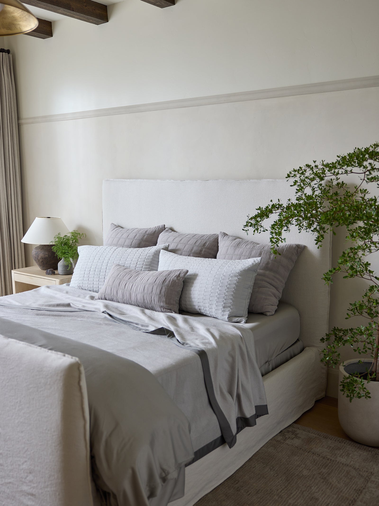 A contemporary bedroom showcases a bed perfectly dressed in gray and white linens, adorned with multiple pillows and a Bamboo Blanket from Cozy Earth. A large potted plant and a nightstand featuring a lamp along with small greenery introduce an element of nature. The room's light-colored walls contribute to its cozy, minimalist aesthetic. 