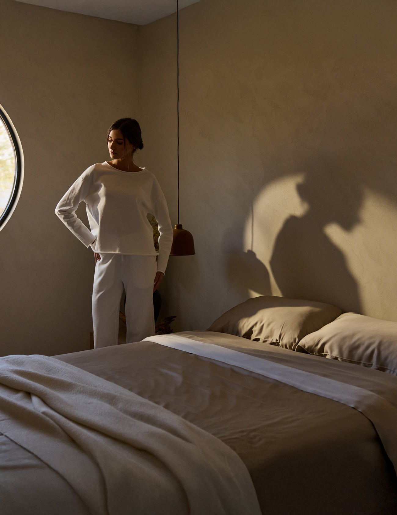 A person stands near a neatly made bed in a softly lit room with beige walls, wearing a white outfit and gazing out a round window. A pendant light casts shadows above the bed, which is adorned with neutral-toned linens featuring Cozy Earth's Bamboo Fitted Sheet. 