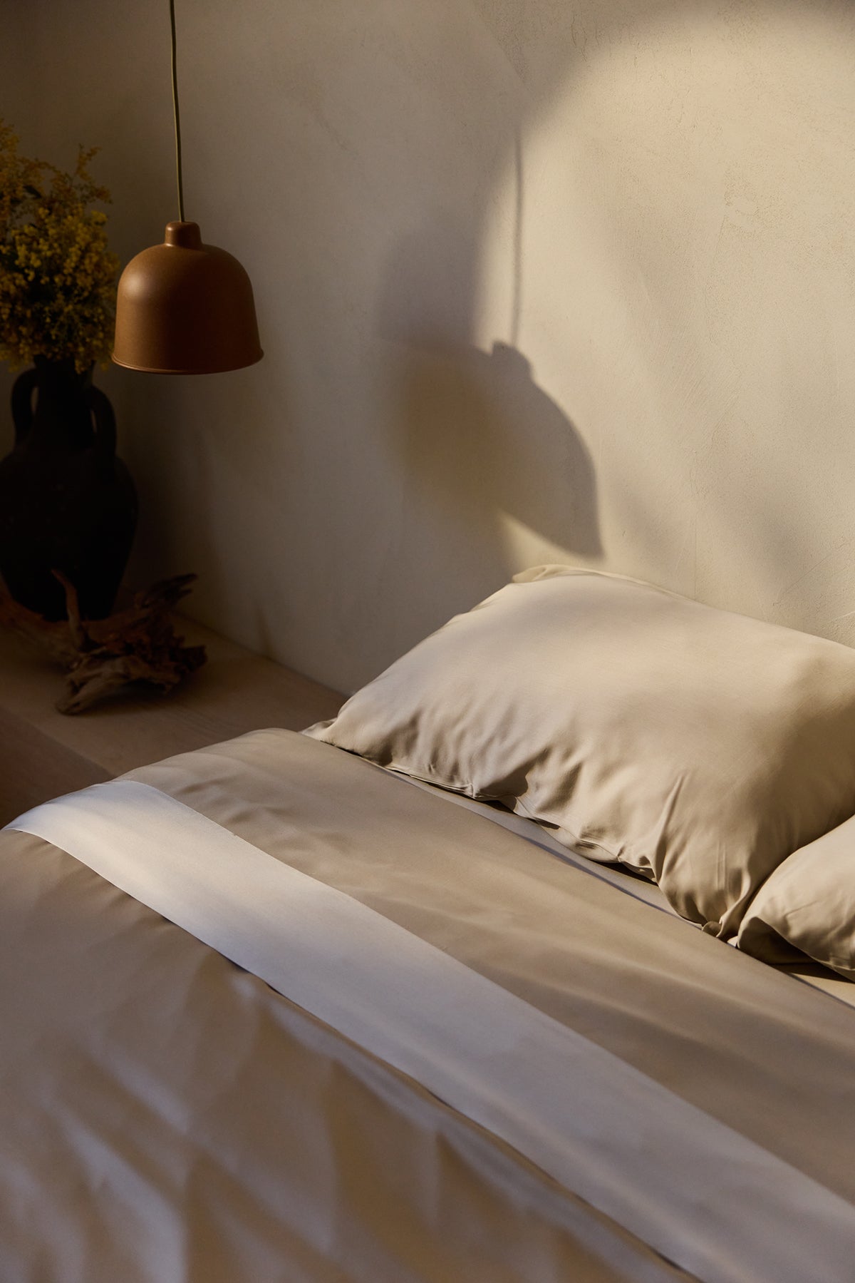 A bed adorned with Cozy Earth's Bamboo Flat Sheets in beige baskes in warm sunlight. A brown hanging lamp and a vase of dried flowers sit on the bedside table, casting gentle shadows on the light-colored wall. 