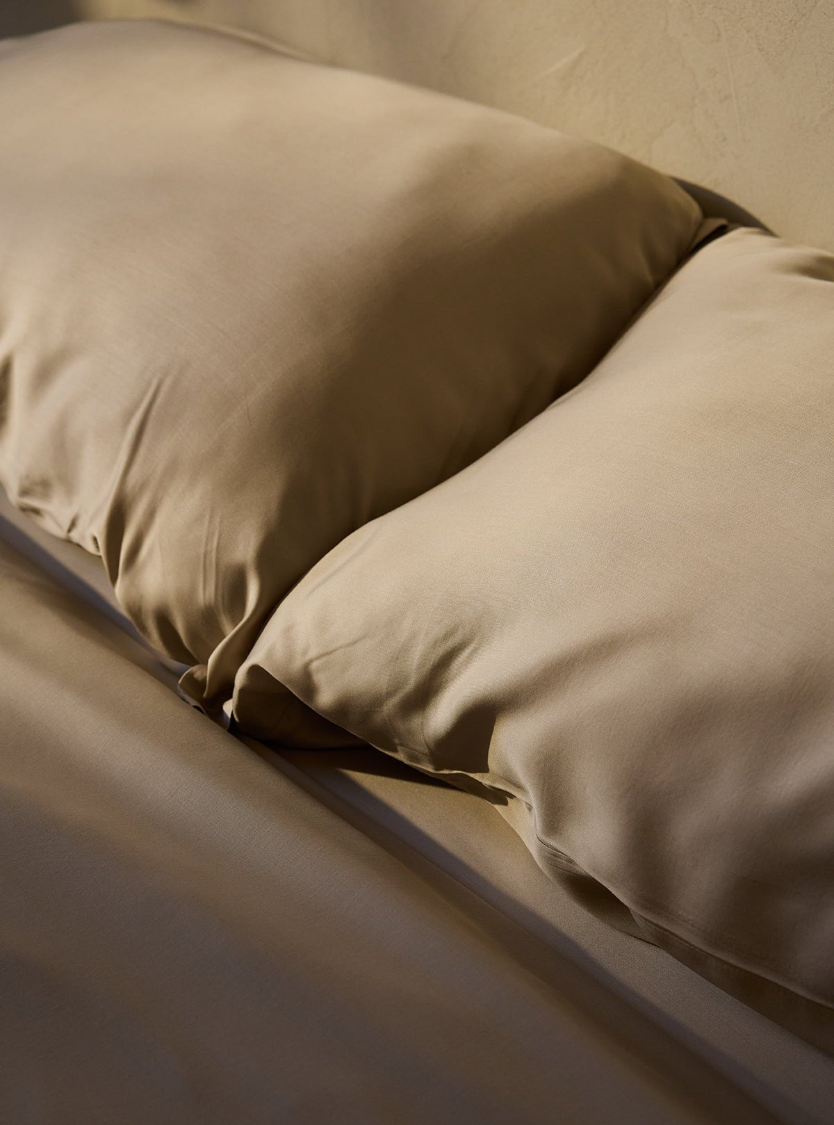 Two Bamboo Pillowcases by Cozy Earth rest closely on a matching bedspread. Ambient lighting highlights their smooth texture and gentle fabric folds, creating a cozy, inviting atmosphere. |Color:Driftwood