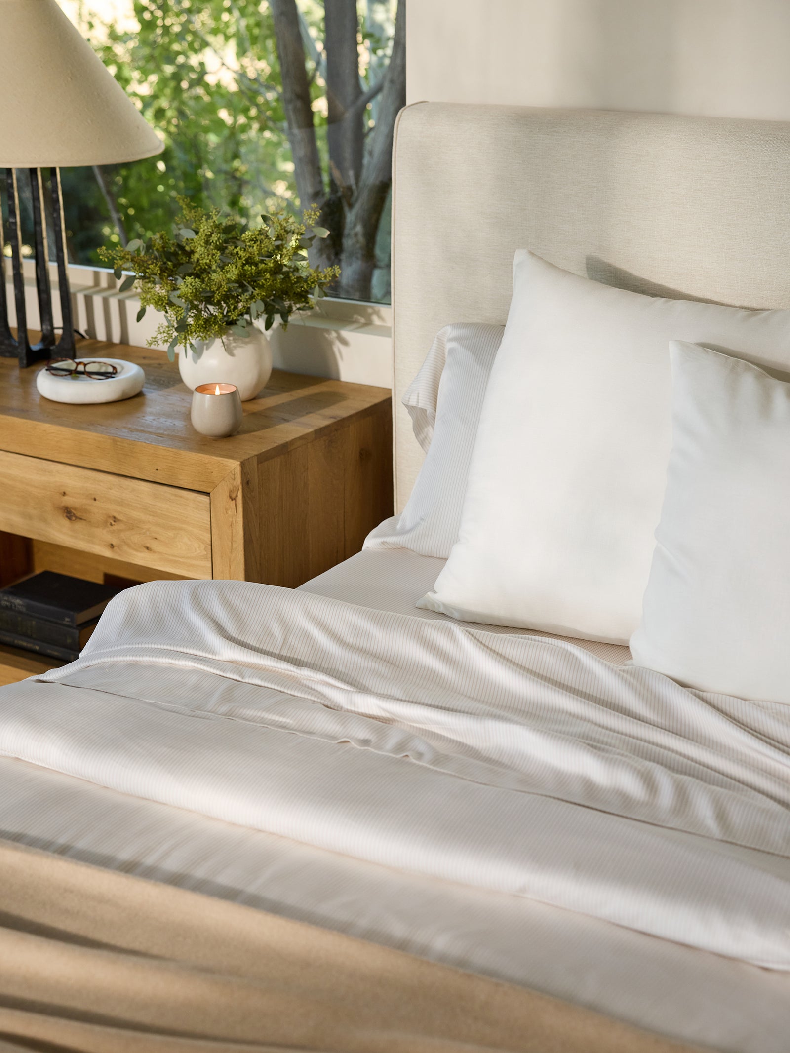 A cozy bedroom scene showcases a neatly made bed featuring Bamboo Pillowcases from Cozy Earth, along with crisp white sheets. A wooden nightstand supports a lamp, a small lit candle, and a vase holding vibrant green foliage. Sunlight flows through the window, casting a warm ambiance throughout the room. 