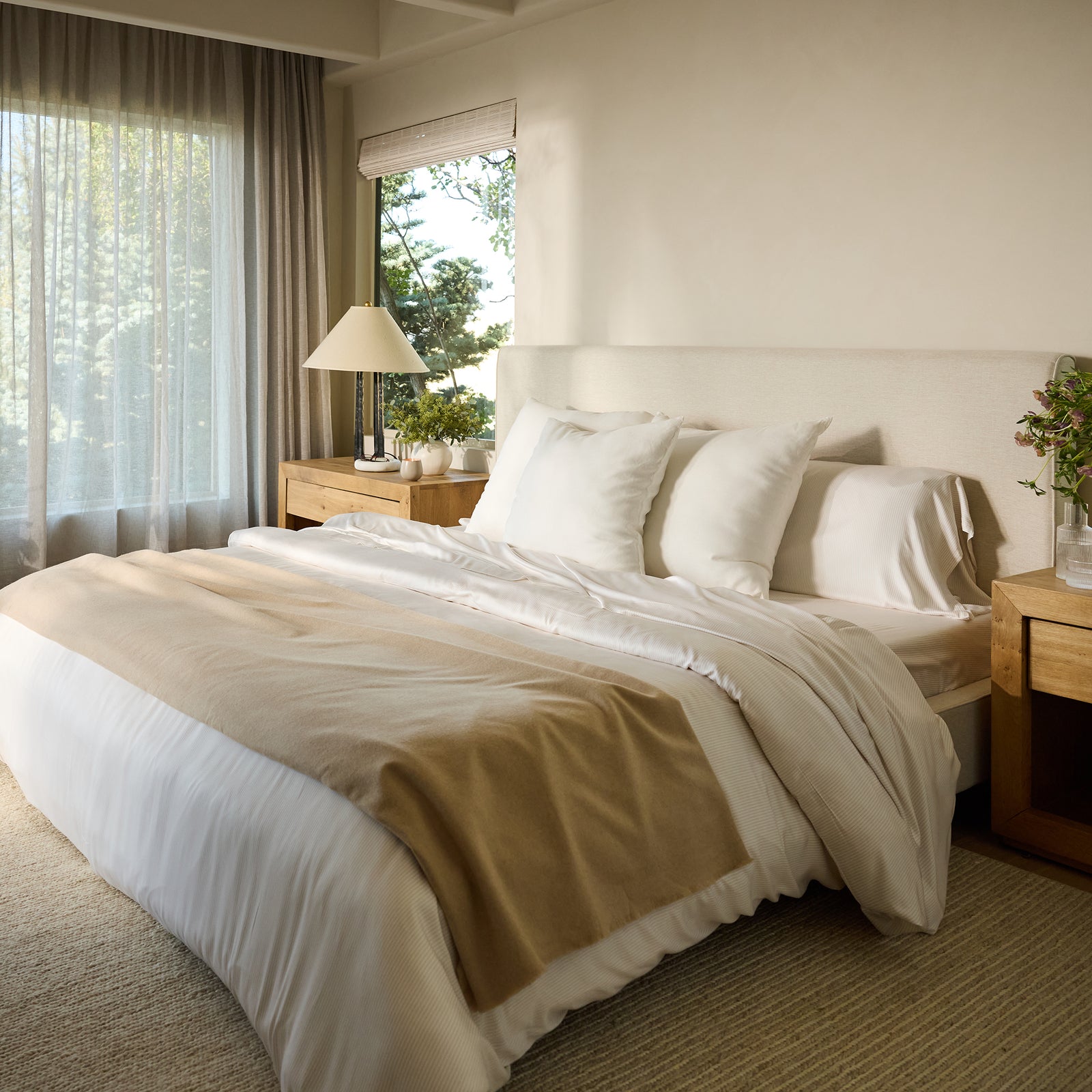 A cozy bedroom showcases a neatly made bed adorned with the Bamboo Sheet Set from Cozy Earth in soothing white and beige tones. Wooden side tables, each decorated with a lamp and flowers, flank the bed. Natural light pours through a large window dressed with sheer curtains, crafting a serene and inviting atmosphere. 