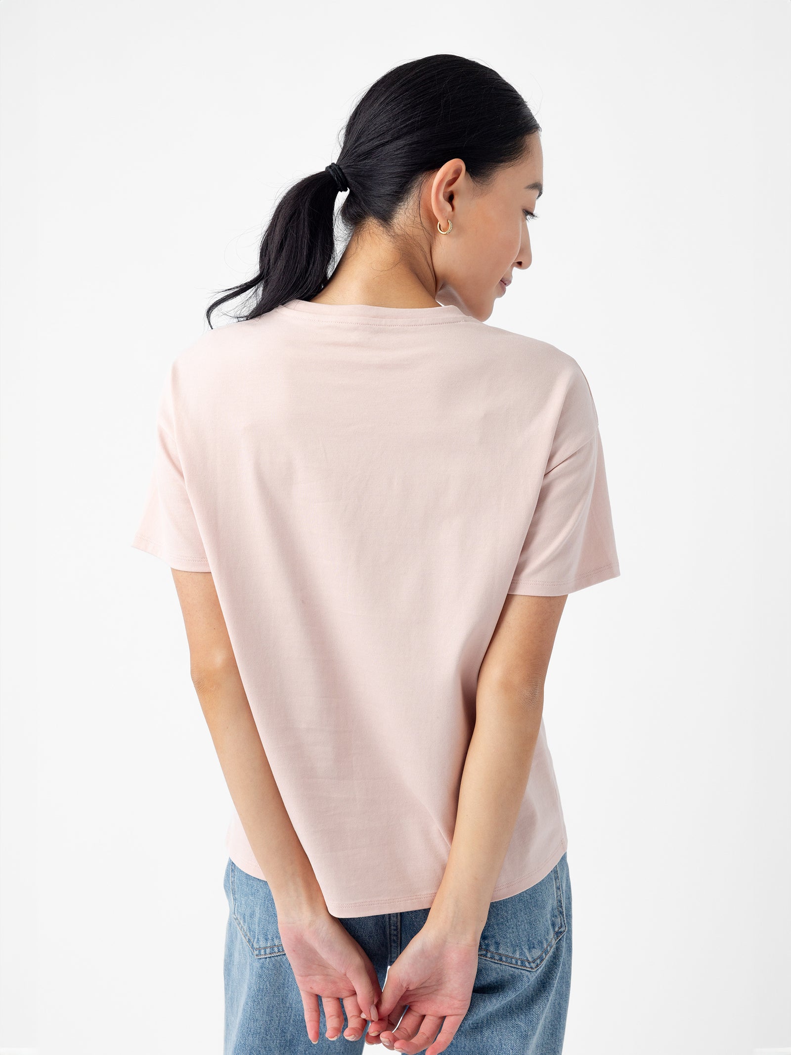 A person with long, dark hair tied in a ponytail is standing with their back to the camera. They are wearing a light pink Women's All Day Tee by Cozy Earth and blue denim jeans. Their hands are loosely clasped behind their back. The background is plain and white. 