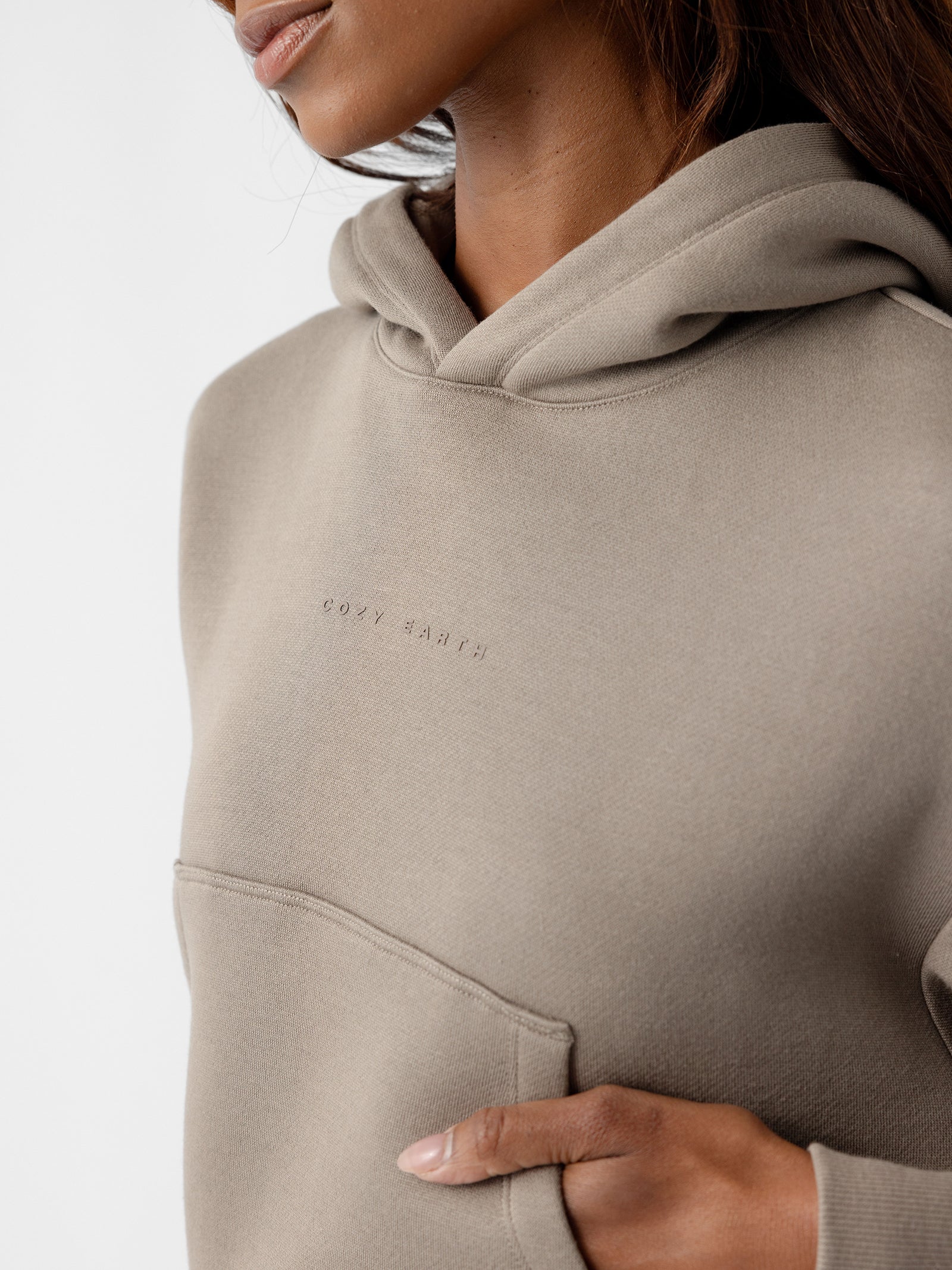 A close-up shot captures a person wearing the Women's CityScape Cropped Hoodie by Cozy Earth. The lower part of their face, neck, and upper torso are shown. This beige hoodie features a front pocket and text embroidered on the chest. The individual’s hand rests inside the pocket, against a plain white background. 