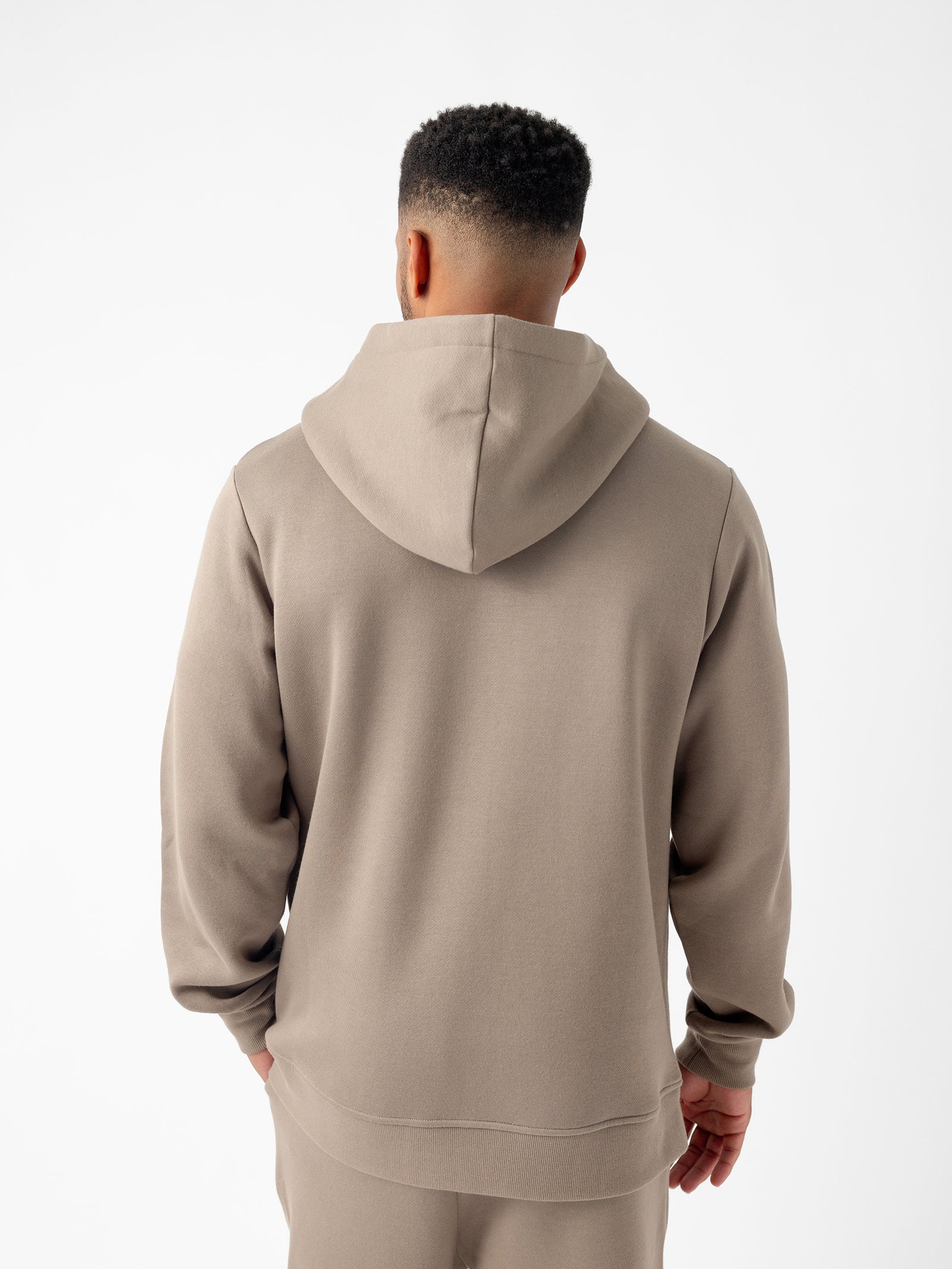 A person stands facing away from the camera, wearing a light brown Men's CityScape Hoodie and matching pants from Cozy Earth. The background is plain white, highlighting the outfit. The hoodie is plain with no visible designs or logos. 