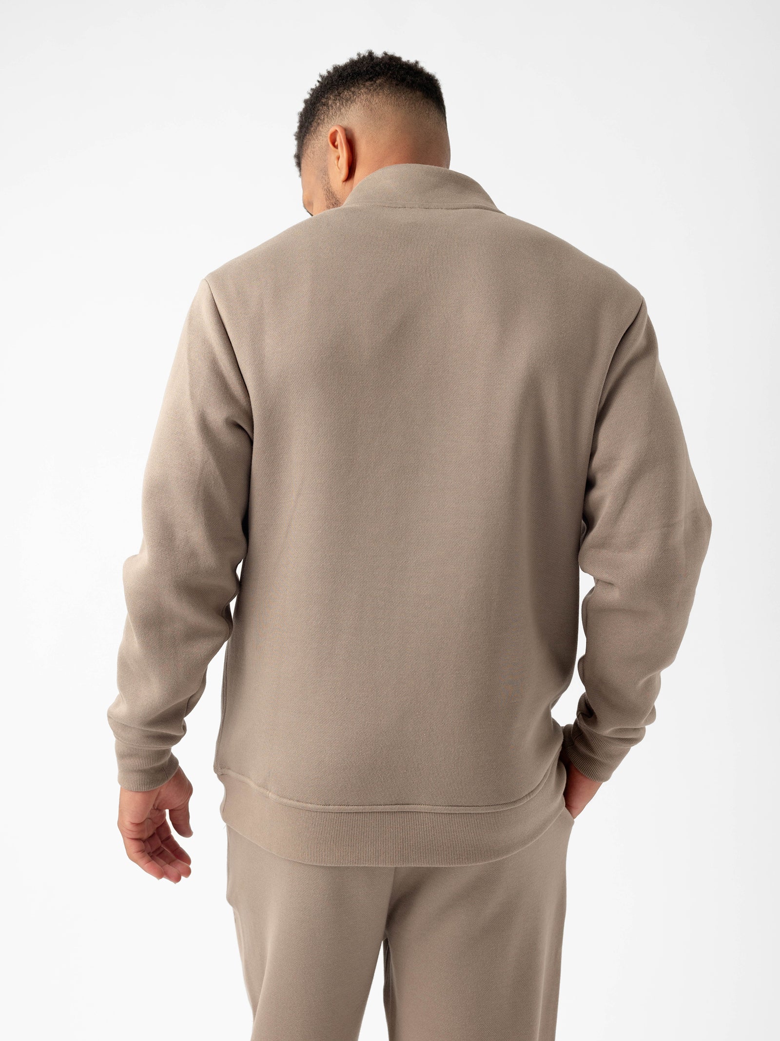 A person with short, curly hair is standing with their back facing the camera, wearing a light brown Cozy Earth Men's CityScape Quarter Zip fleece sweater and matching pants. The background is plain white. 