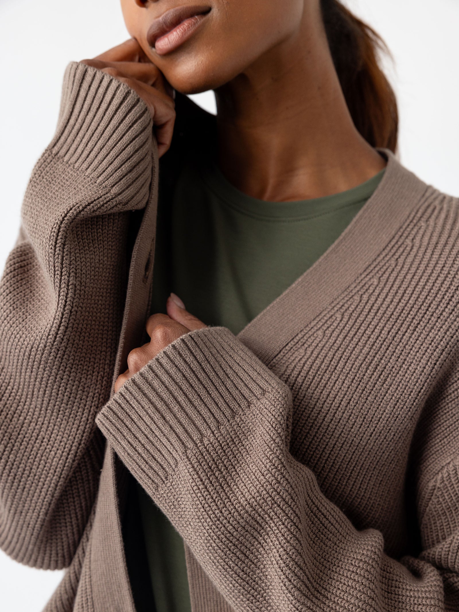 A person wearing the Oversized Classic Cardigan by Cozy Earth in taupe and a green shirt, partially covering their face with the cardigan's collar. The image highlights the texture of the sweater and showcases the person's serene expression. 