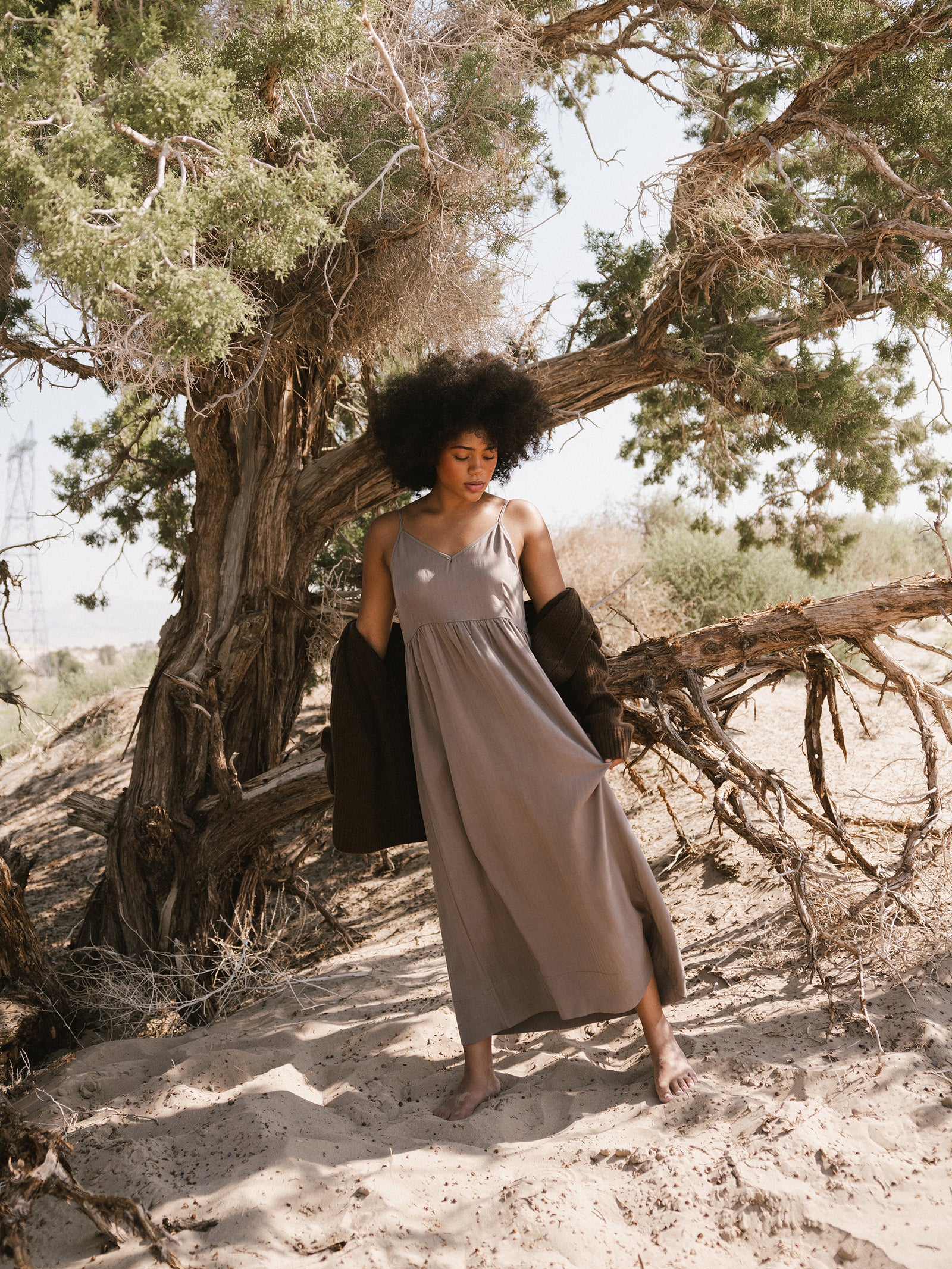 A woman with curly hair stands barefoot on sandy ground, wearing the Women's Sunset Gathered Dress by Cozy Earth in a long, light brown shade and a dark jacket draped off her shoulders. She is positioned under a large, gnarled tree with green foliage in a desert-like setting. 