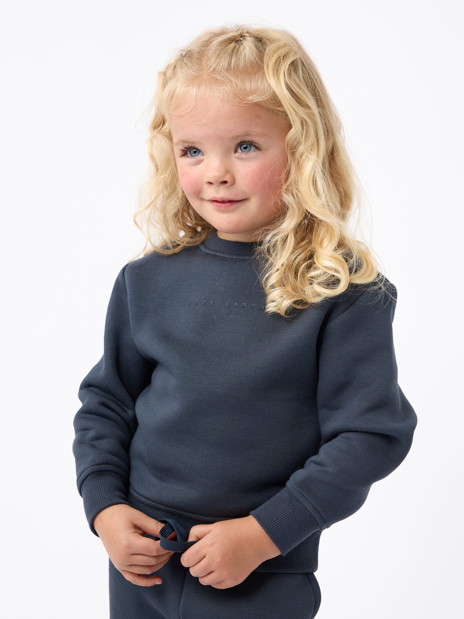 A young child with curly blonde hair is wearing Cozy Earth's Kid's CityScape Crewneck and looking slightly to the side against a plain white background. 