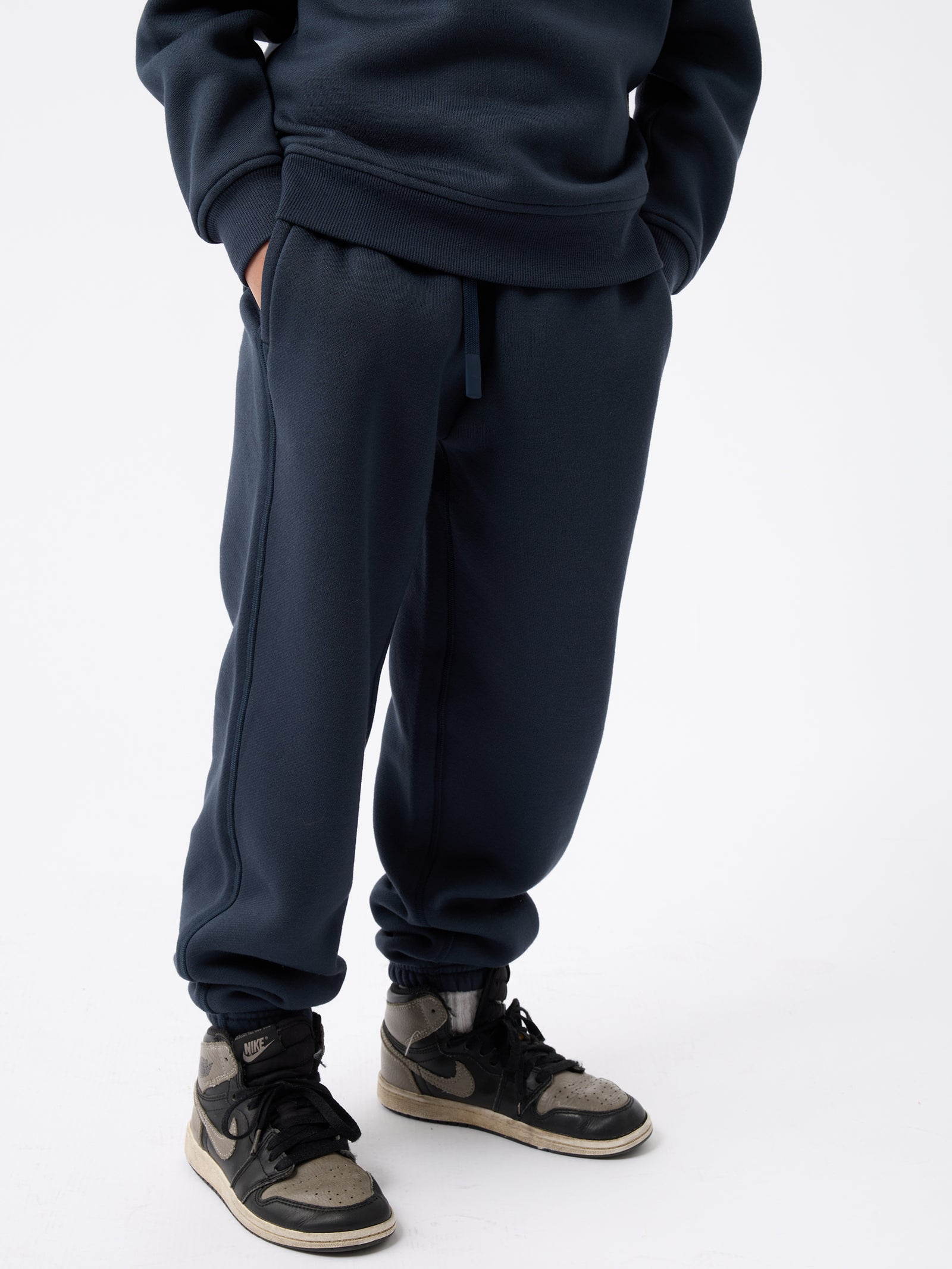 A person wearing Cozy Earth's Kid's CityScape Sweatpants in dark blue, along with a matching sweatshirt, stands against a white background. They are also wearing high-top sneakers in black and gray. The faces and upper body are not visible. 