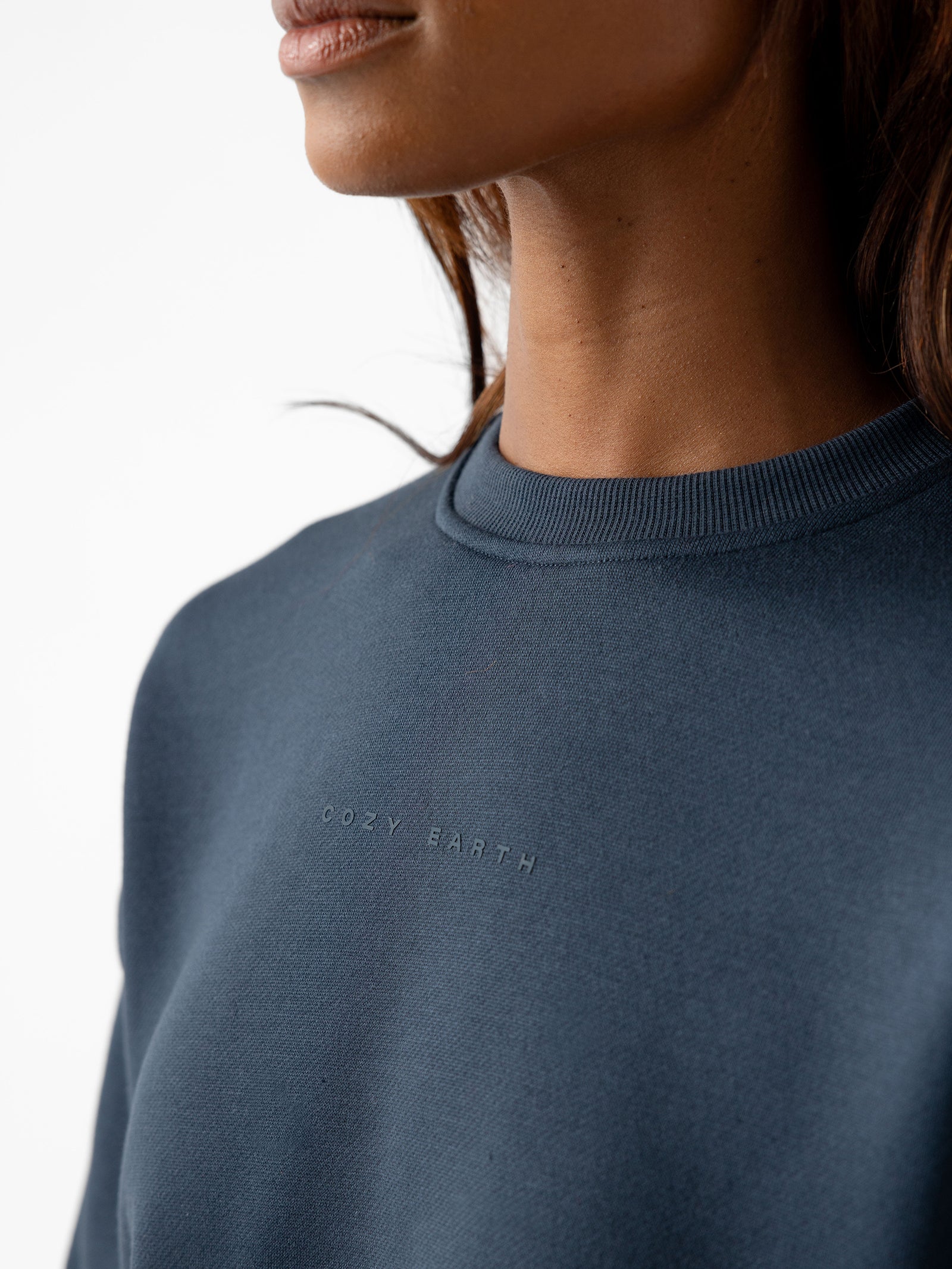 Close-up of a person wearing the Women's CityScape Crewneck in dark blue, featuring the text "COZY EARTH" embroidered in small letters on the chest. Only the lower part of the person's face and neck are visible, along with some dark hair falling to the side. 