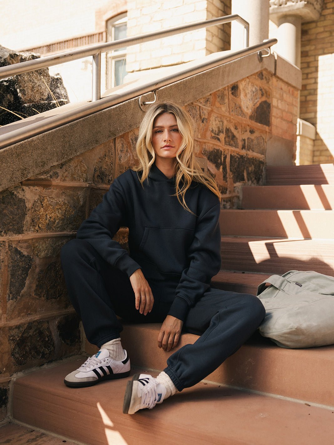 A person with long, blonde hair sits on outdoor steps made of brick and concrete. They are wearing a dark Women's CityScape Cropped Hoodie from Cozy Earth and matching sweatpants, paired with white Adidas sneakers. A grey backpack is placed beside them. The person has a serious expression. 