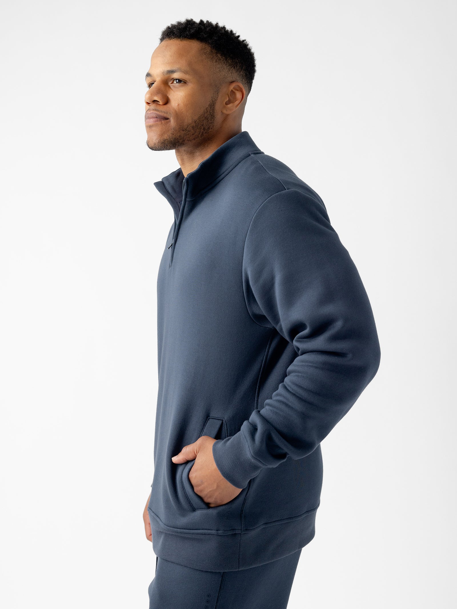 A man with short hair is standing against a plain white background, wearing a Cozy Earth Men's CityScape Quarter Zip in navy blue. He has his left hand in his pocket and is looking to the side. 