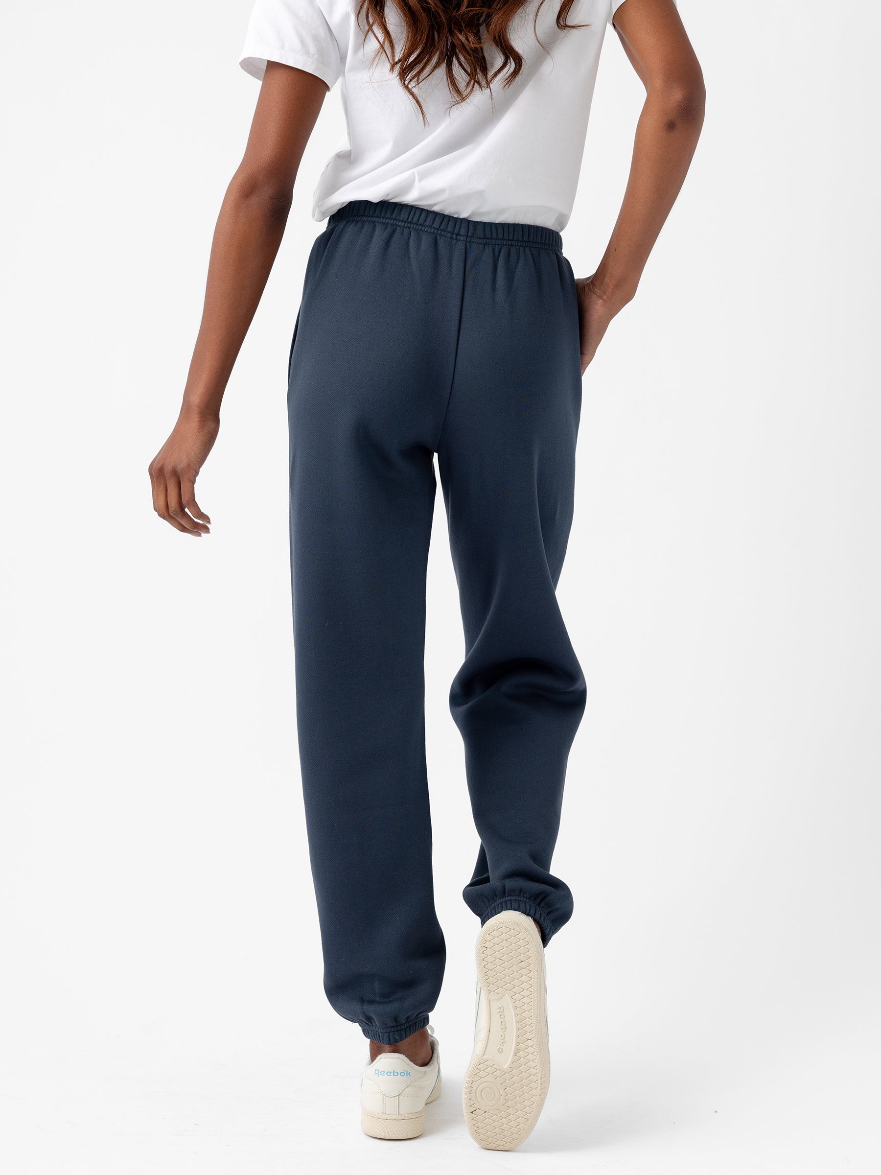 A person with long hair is shown from the back wearing a white short-sleeve shirt and navy blue Women's CityScape Sweatpant by Cozy Earth. They are also wearing white sneakers. The background is plain white. |Color:Eclipse