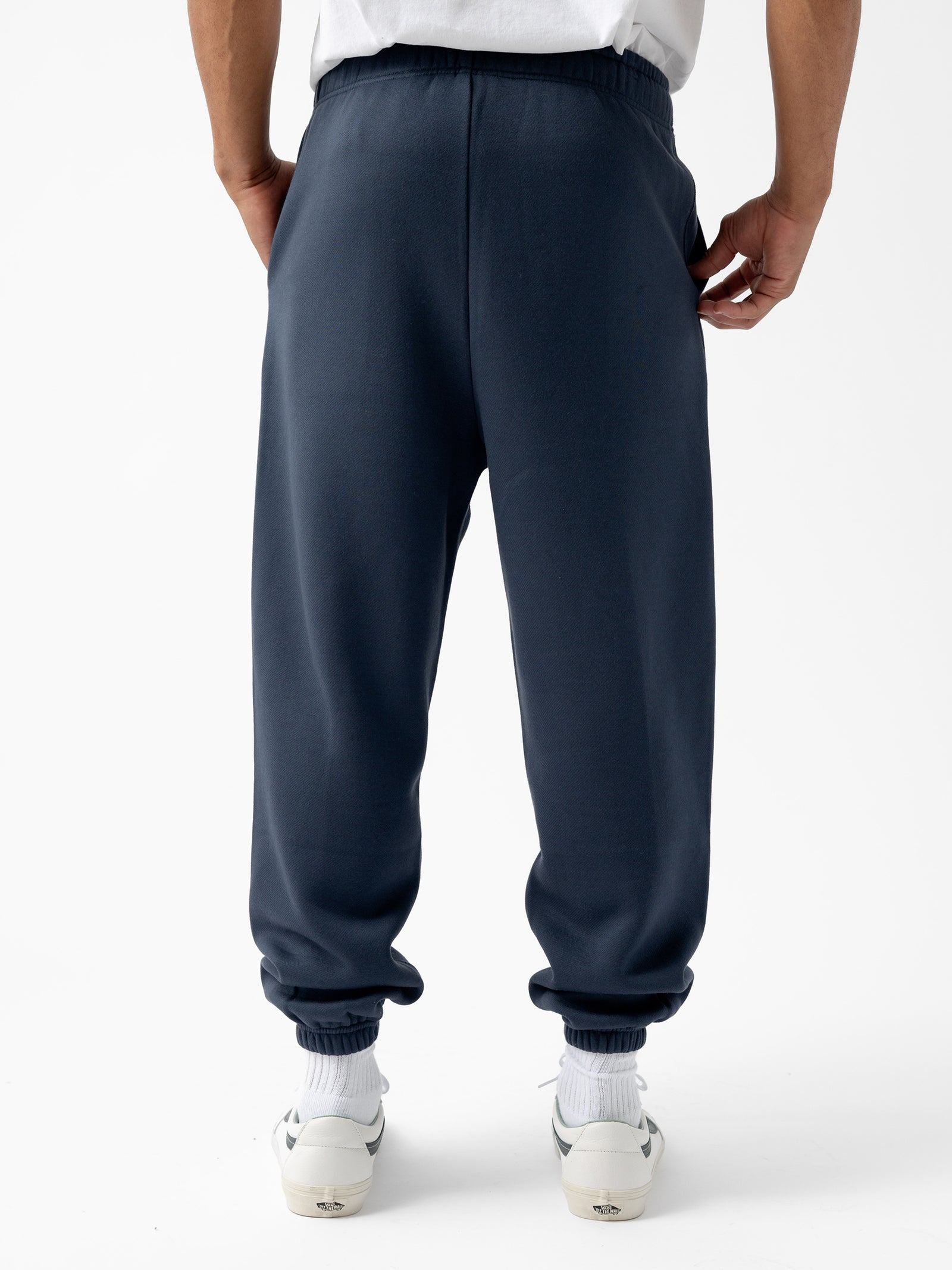 A person wearing the Men's CityScape Sweatpant by Cozy Earth in dark blue and white sneakers stands facing away, hands in pockets. The sweatpants feature elastic cuffs at the ankles. The background is plain white. 