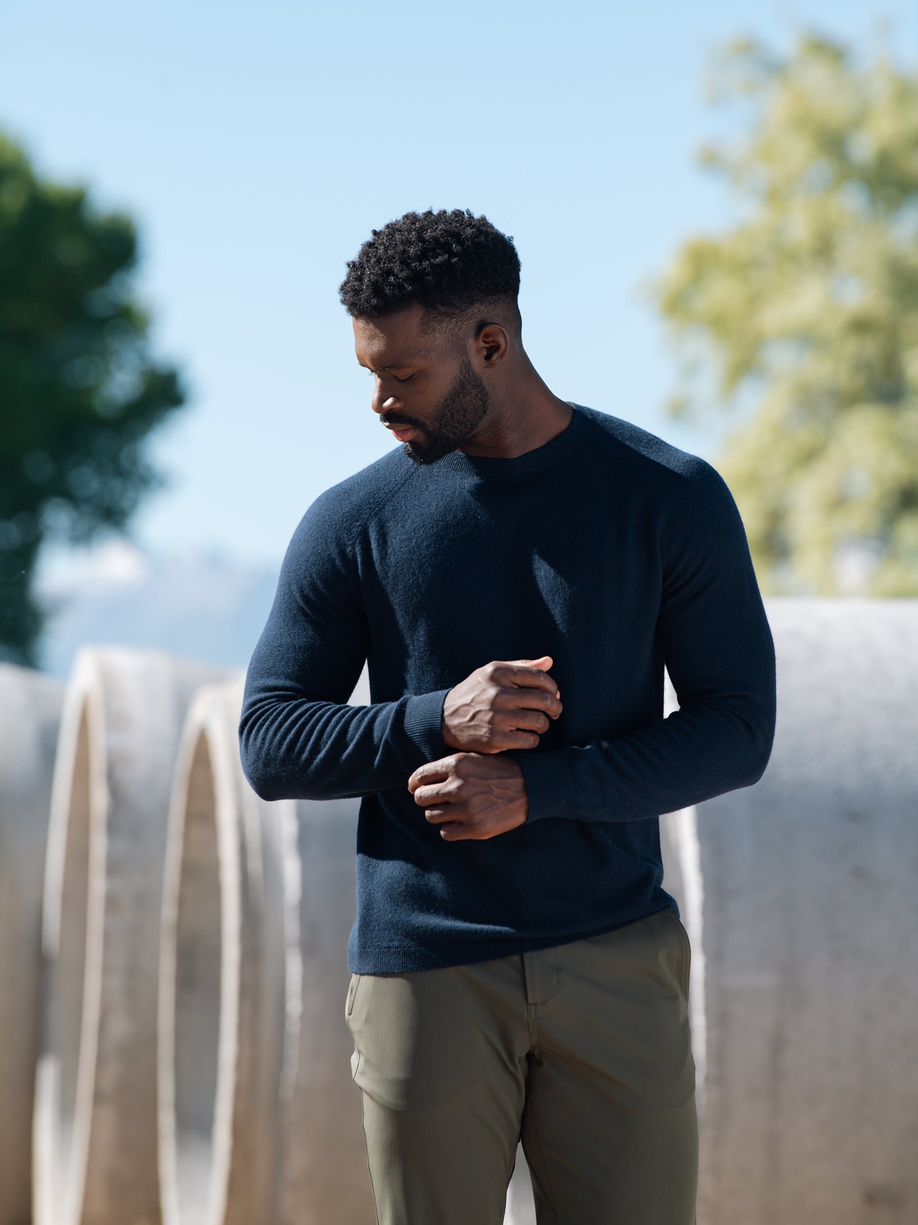A man stands outdoors facing slightly downward. He is wearing a dark blue Cozy Earth Men's Crewneck Sweater and light brown pants. In the background, there are large concrete pipes and trees under a clear blue sky. 