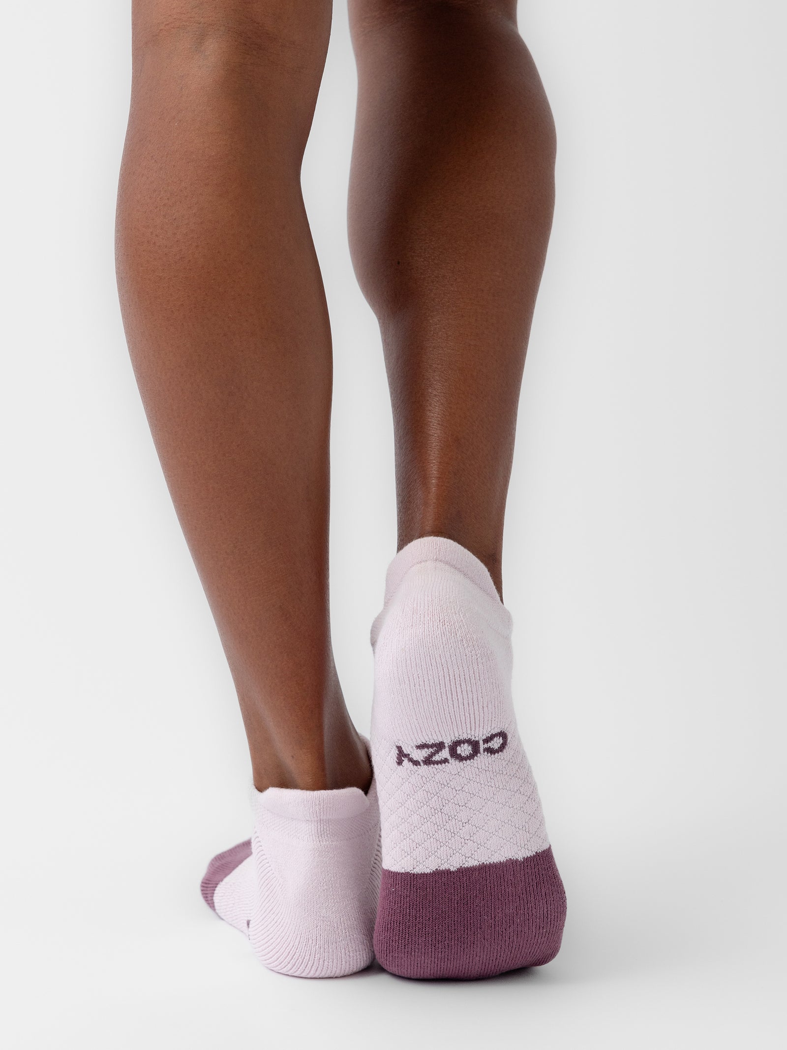 A close-up shot showcases a person's lower legs and feet adorned with white ankle socks that feature purple heels and toes. One sock from the Essential Ankle Sock 2-Pack by Cozy Earth displays the word "COZY" in purple text on the back. The background is a plain white, emphasizing the design details of the socks. 