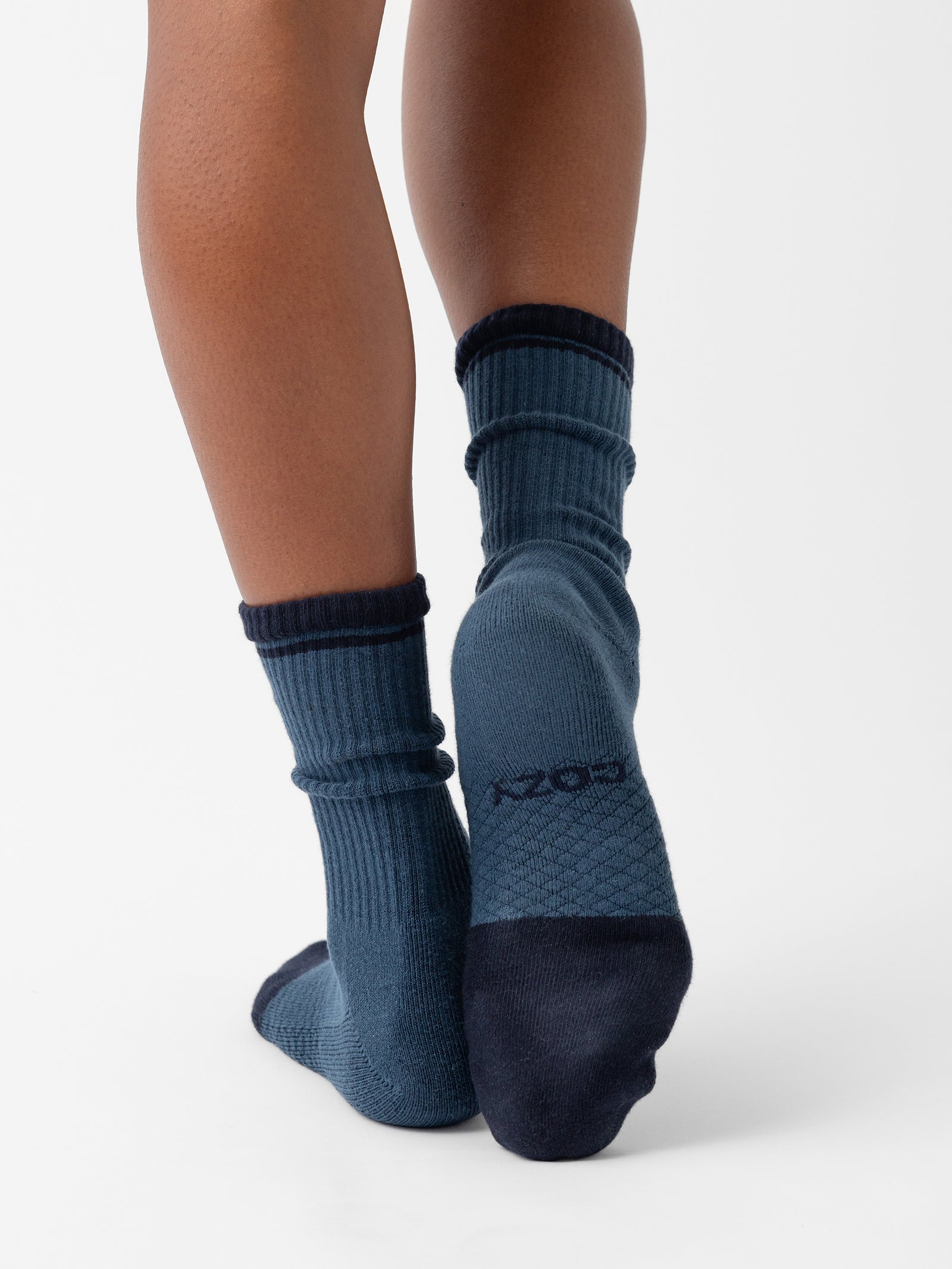 A person wearing a pair of blue Essential Calf Socks with black toe and heel sections from Cozy Earth is standing on a white background. The socks feature ribbed cuffs and a textured design on the soles, with the word "Cozy" partially visible on the sole of one sock. 