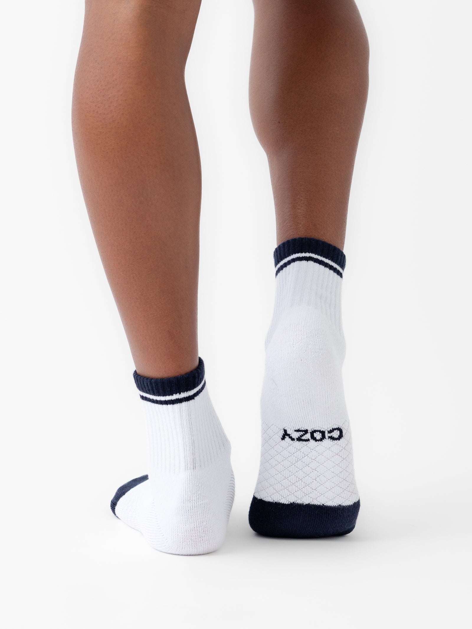 Back view of a person wearing white Essential Quarter Socks with dark blue accents at the cuff, heel, and toe. The right sock features the word "COZY" written on the sole. The person's legs are positioned against a plain, white background. These socks are part of Cozy Earth's Essential Quarter Sock 2-Pack. 