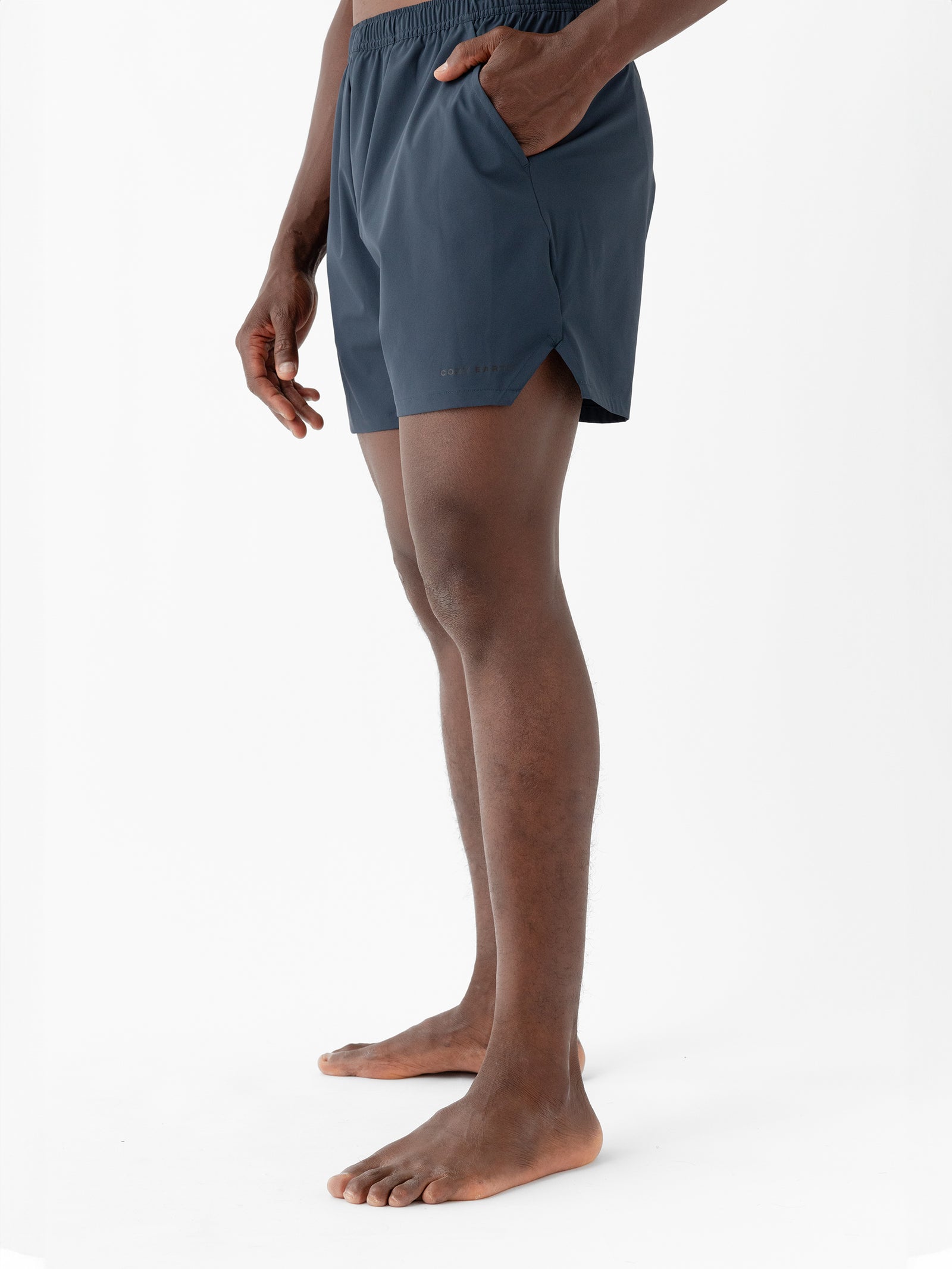 A person stands barefoot against a white background, wearing dark blue Men's Performance Sleep Shorts by Cozy Earth. Their hands are relaxed by their sides, with the right hand placed inside a pocket. The shorts feature a subtle logo near the bottom hem on the left side. 